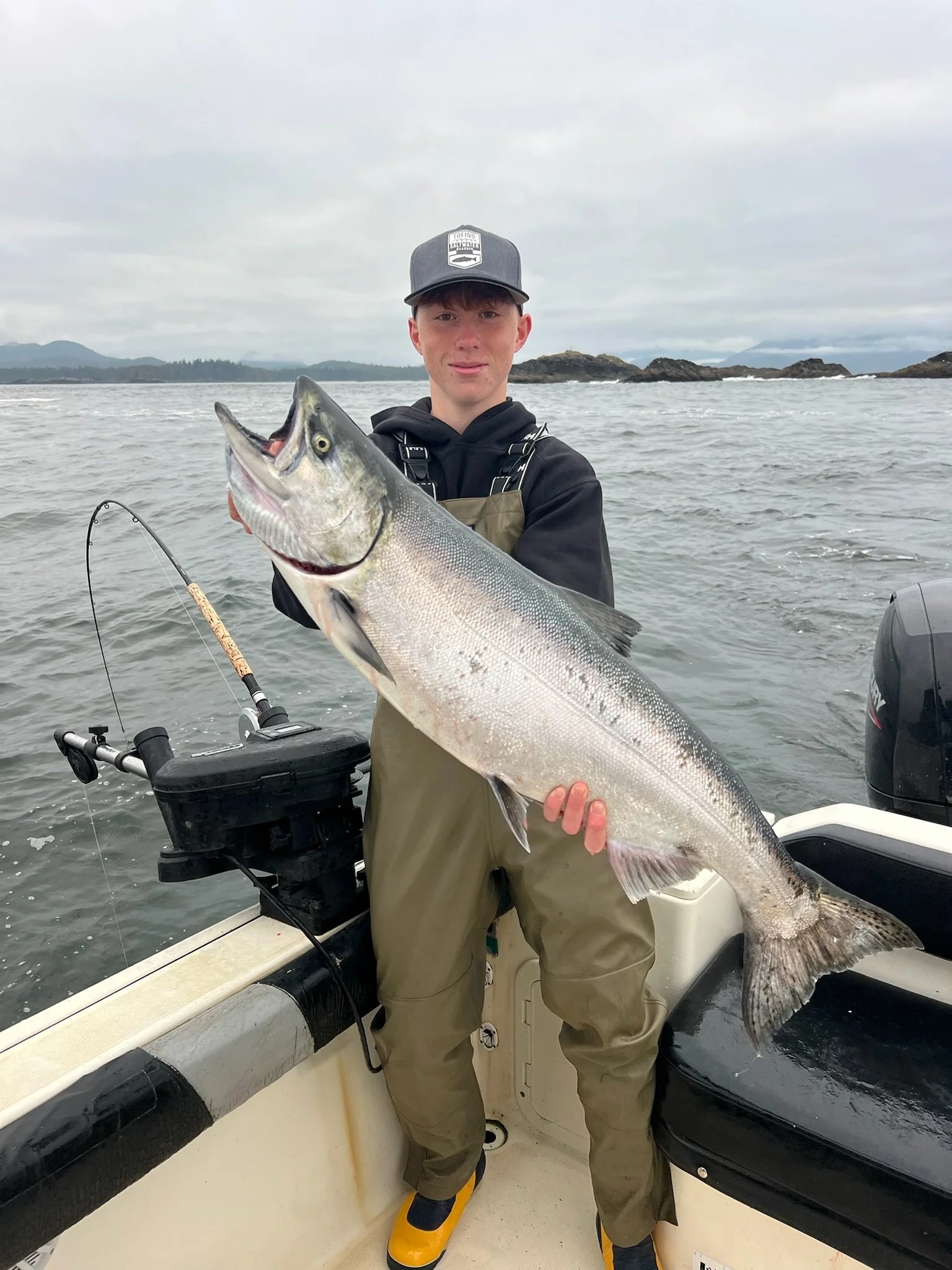 A young man in fishing gear holding a large fish on a boat with water and rocky islands in the background.