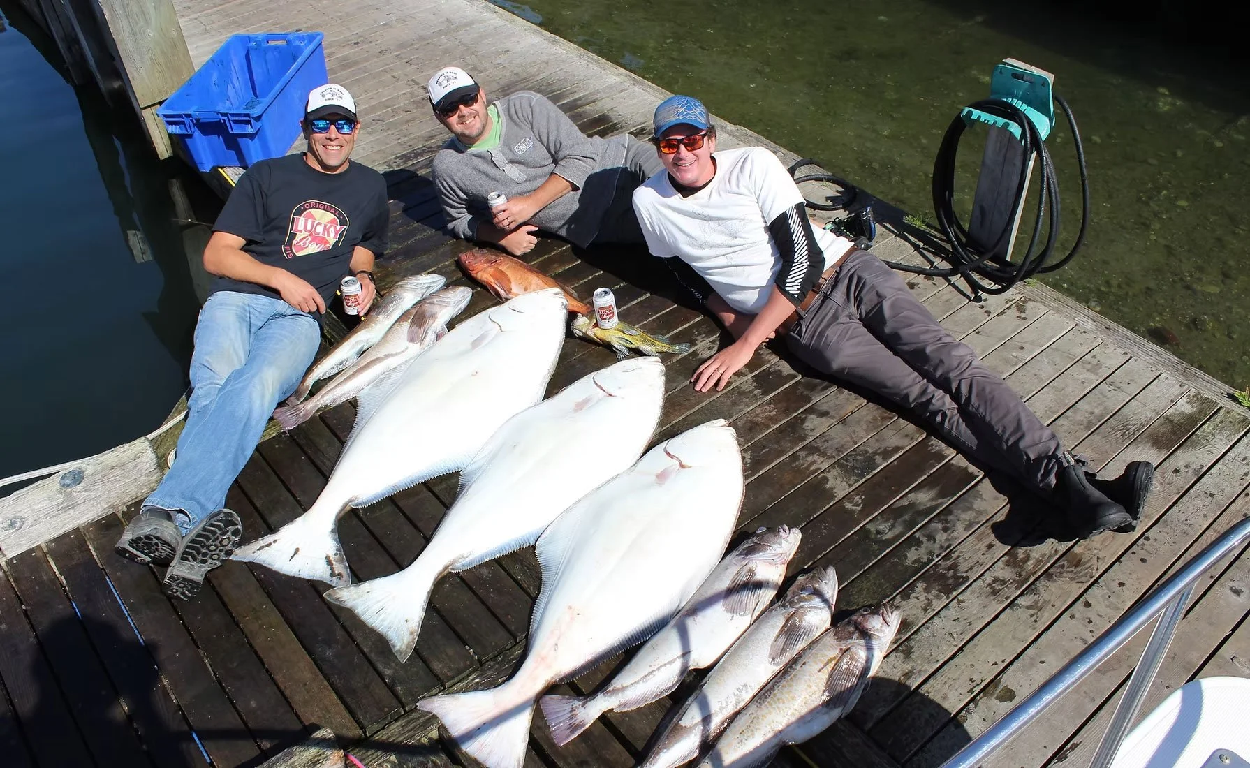 Three men lying on a wooden dock next to each other, smiling, with fish they caught arranged in front of them. Two men are holding cans of beer, and the dock is near water with fishing equipment and a blue tackle box nearby.