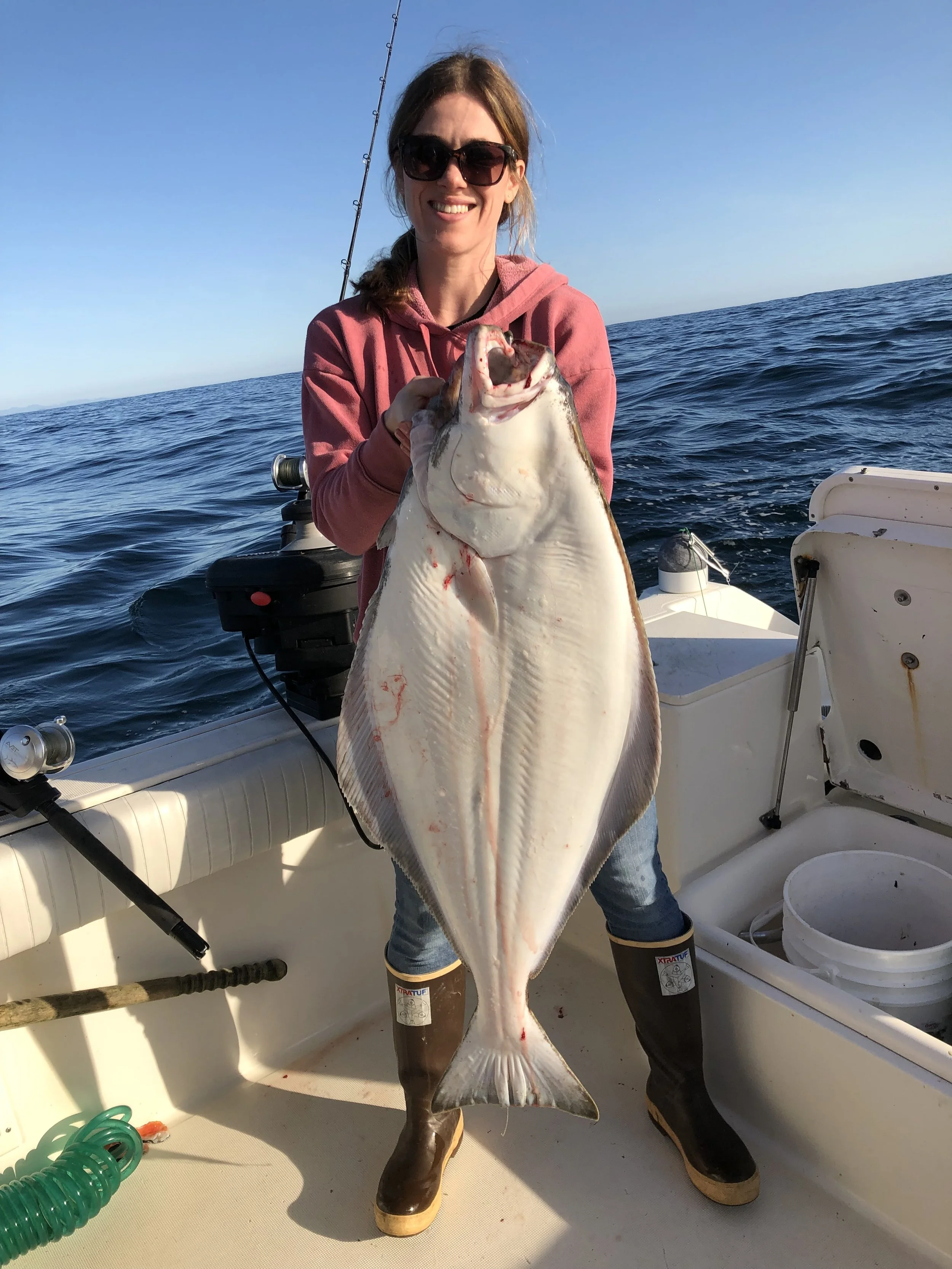 Woman holding a large flatfish on a boat at sea, with fishing gear visible in the background, during daytime.