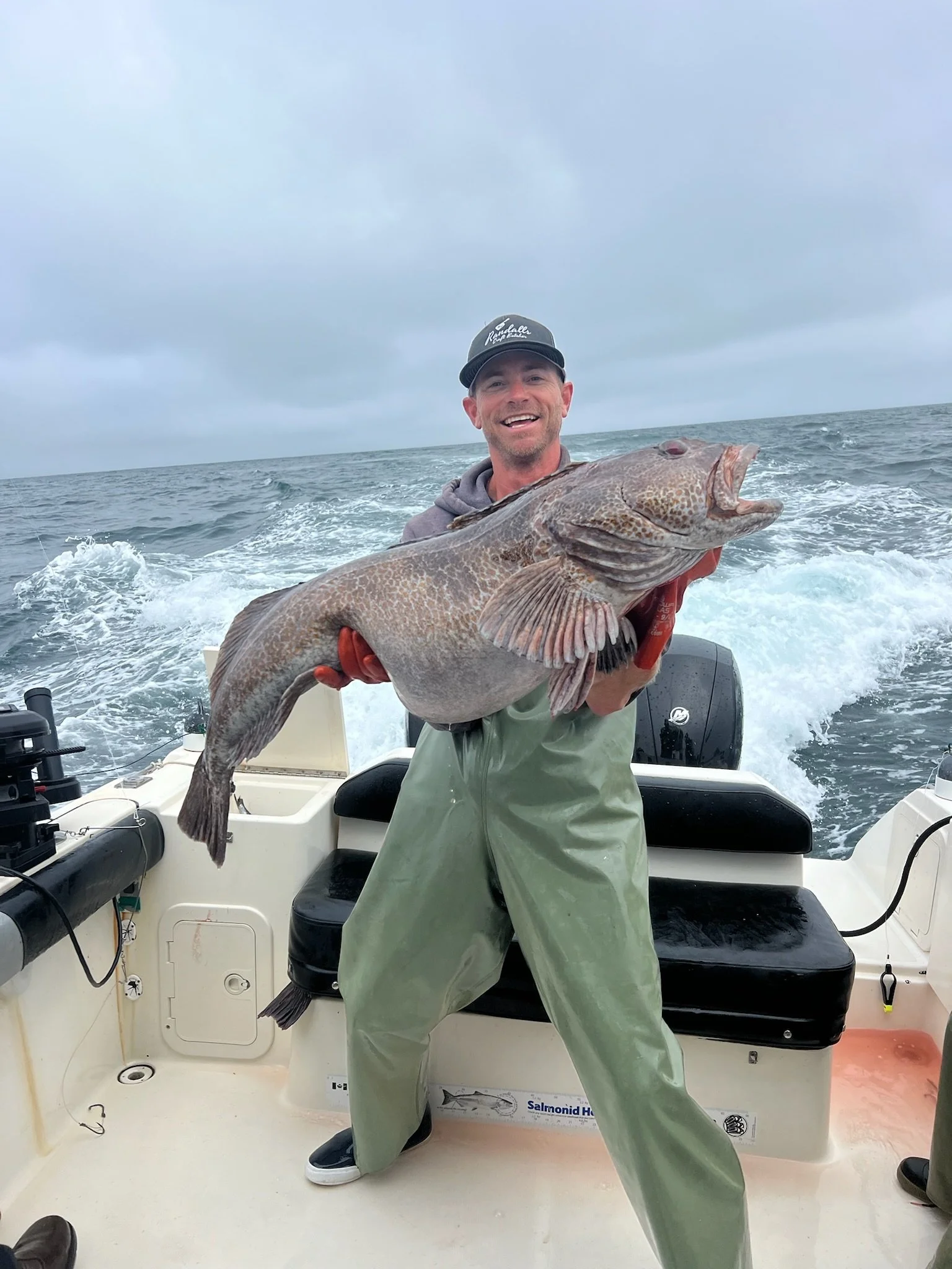 A man wearing a black cap and green waders holding a large fish on a boat in rough ocean water under cloudy sky.