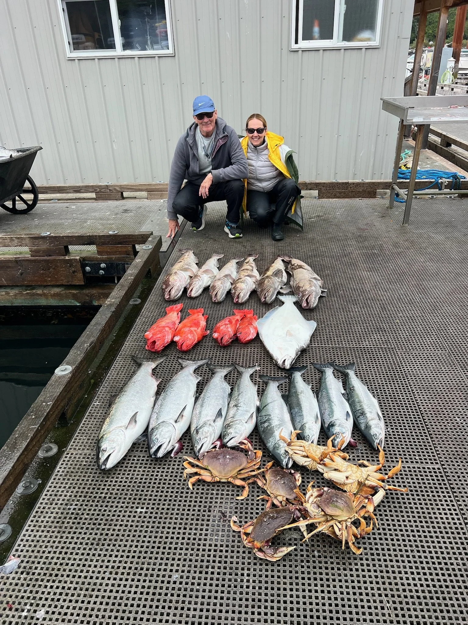 Kneeling man and woman with big smiles posing on a dock, displaying caught fish including salmon, flatfish, and crabs, in front of a building with windows.