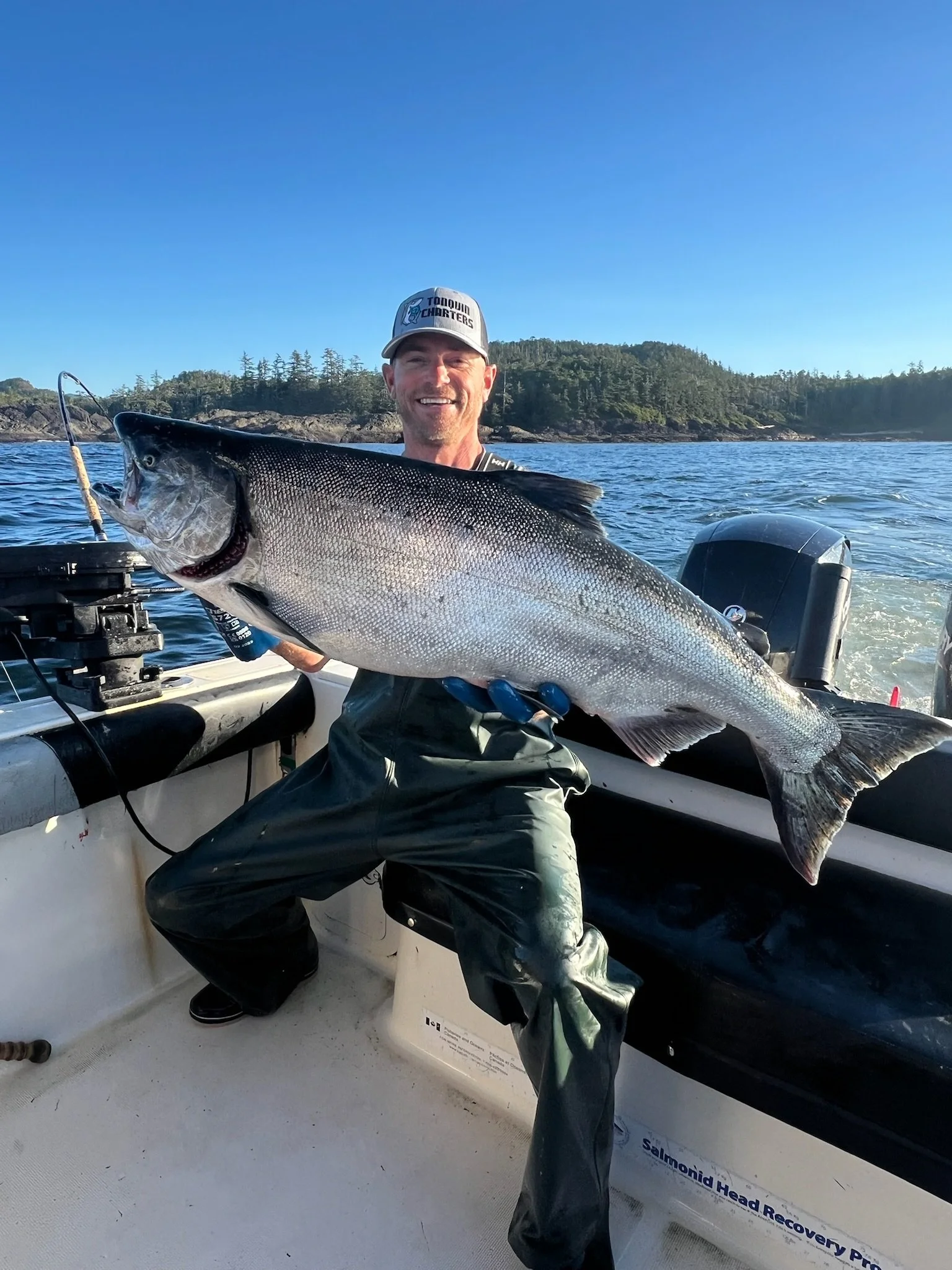 Man on a boat holding a large fish, wearing a gray cap and waterproof clothing, with a scenic water and forested shoreline in the background under clear blue skies.