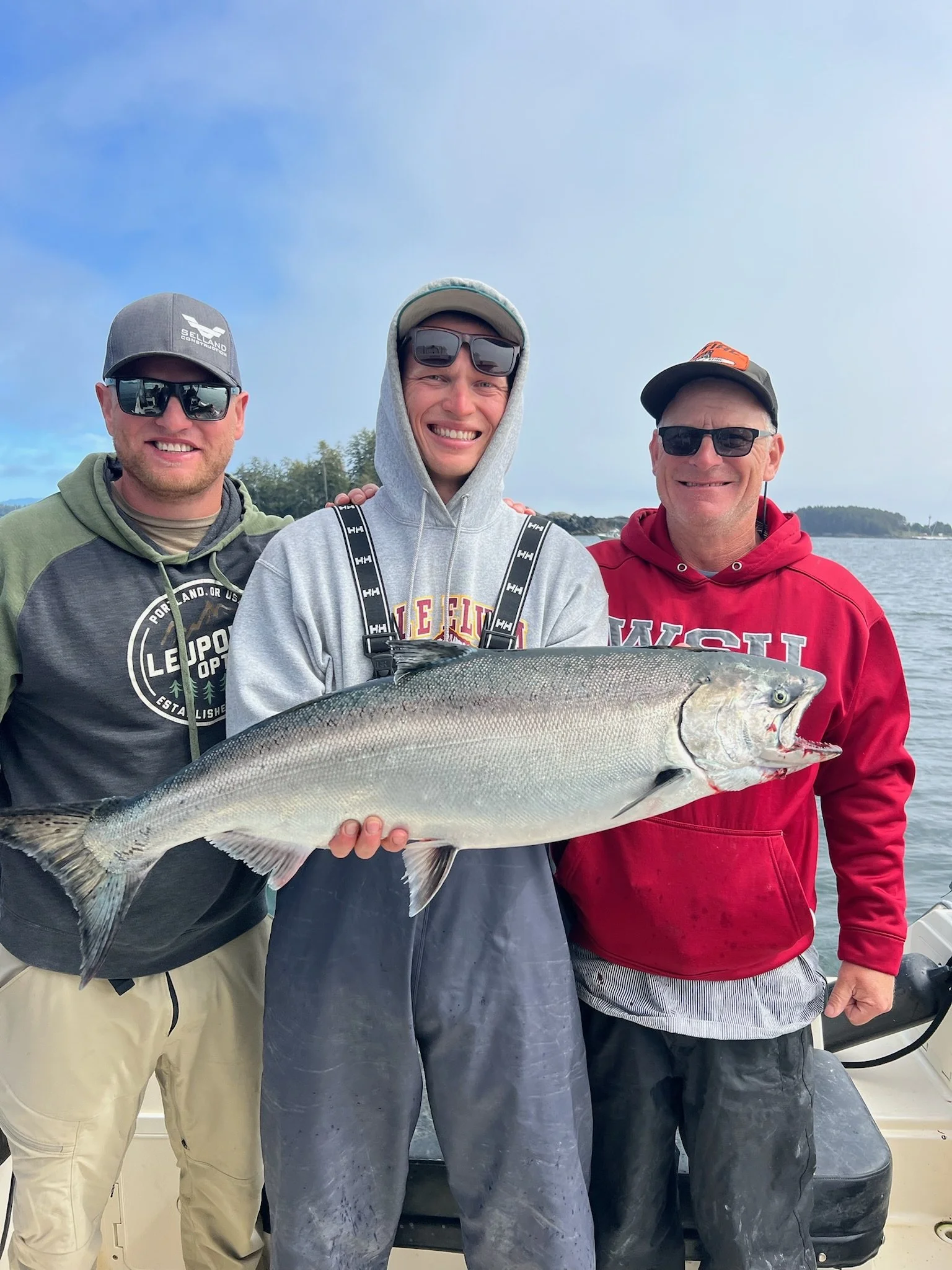 Three men on a boat holding a large fish, smiling at the camera with water and a shoreline in the background.