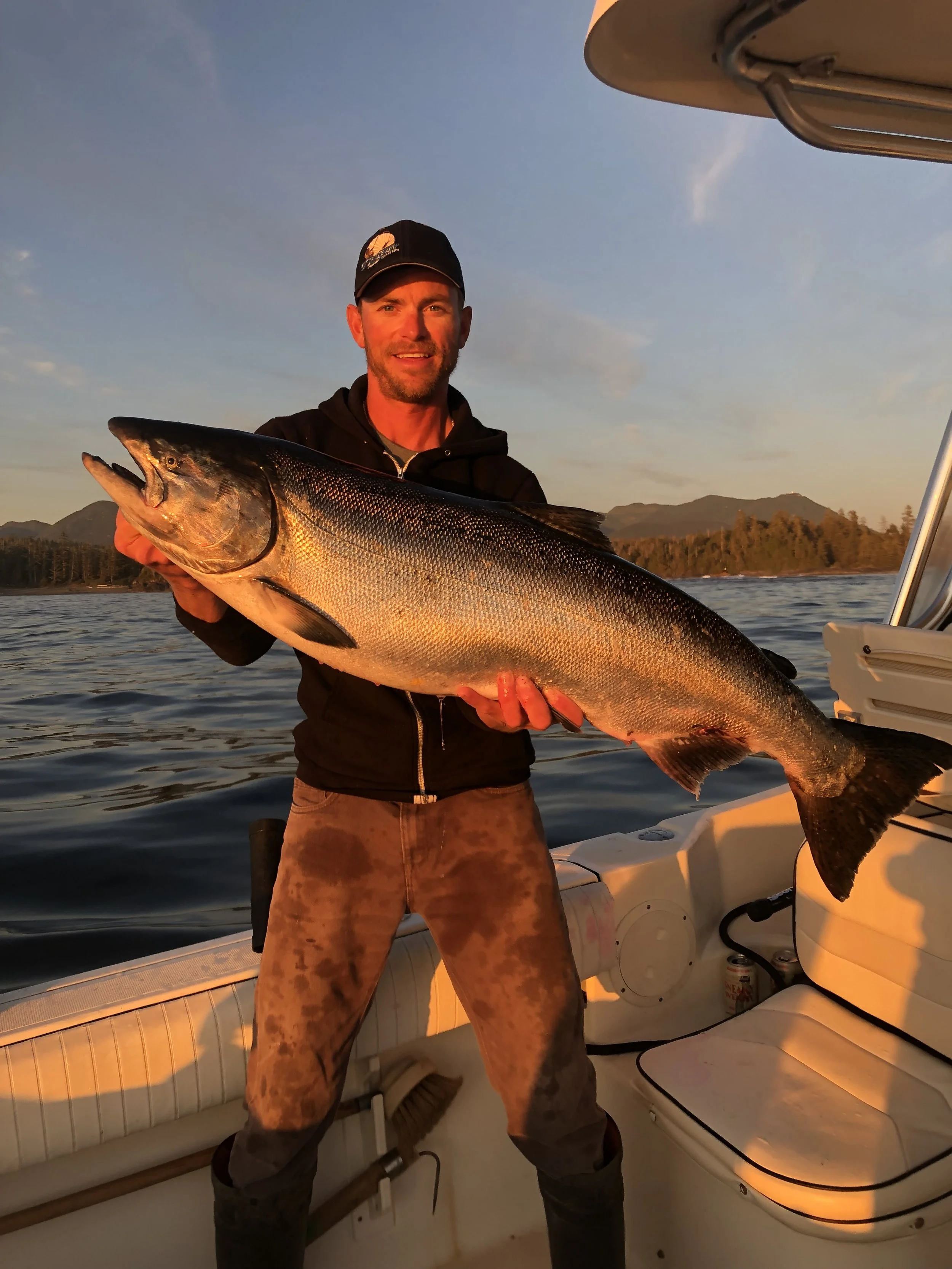 A man holding a large fish on a boat during sunset, with mountains and water in the background.