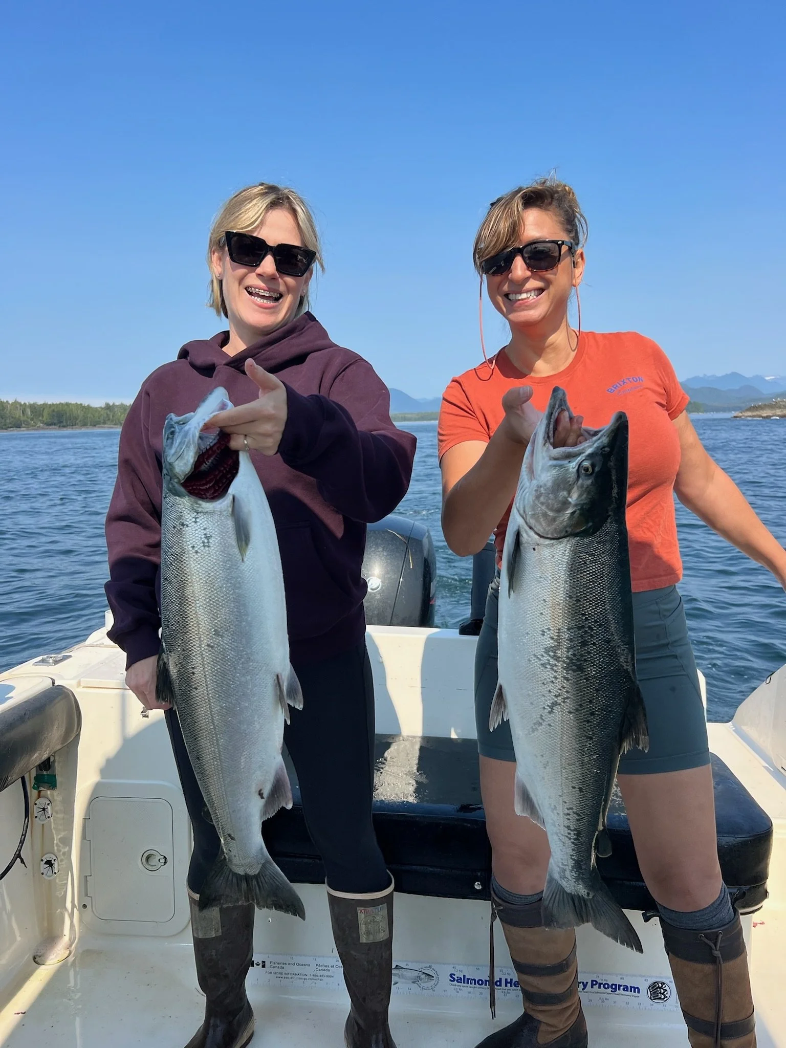 Two women on a boat holding large fish they caught, with a body of water and mountains in the background, wearing sunglasses and casual clothing.