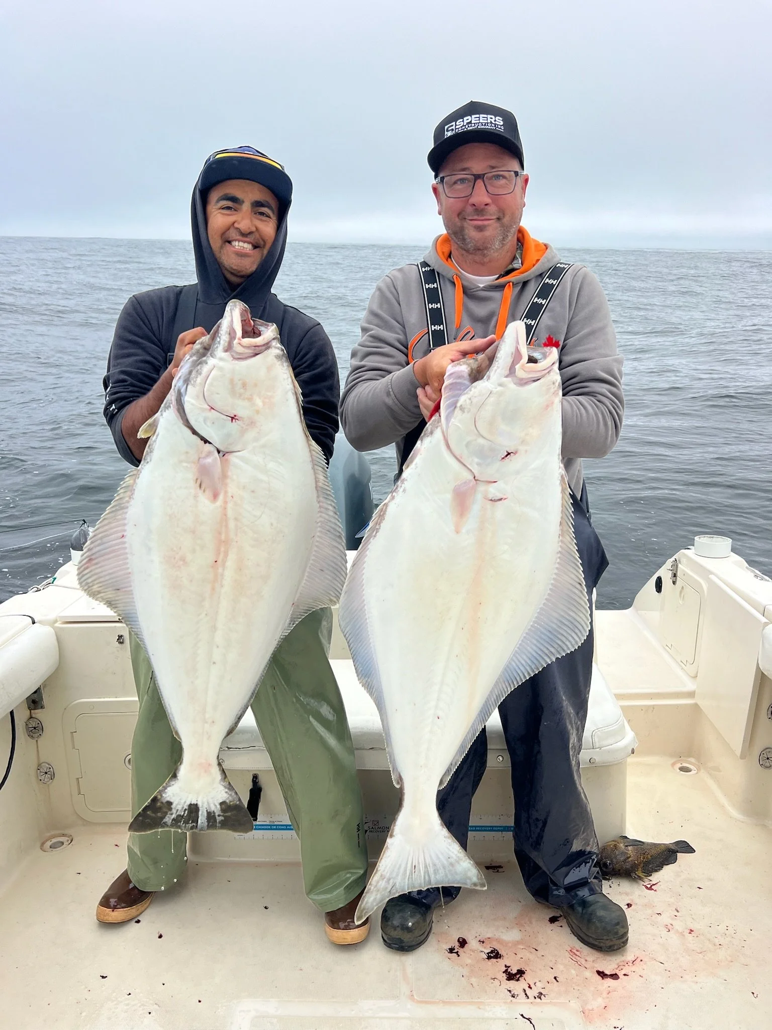 Two men on a boat holding large halibut fish, smiling at the camera with the sea and overcast sky in the background.