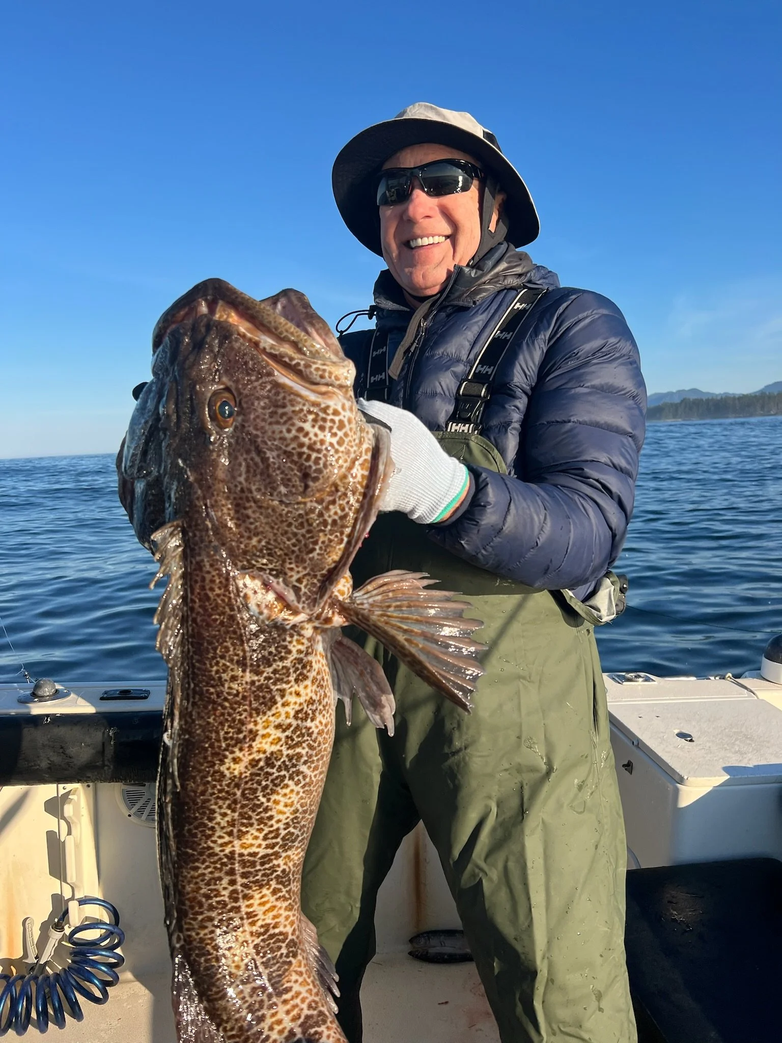 A man smiling and holding a large fish on a boat, wearing sunglasses, a wide-brimmed hat, a dark jacket, and green fishing waders with a backdrop of water and distant land.