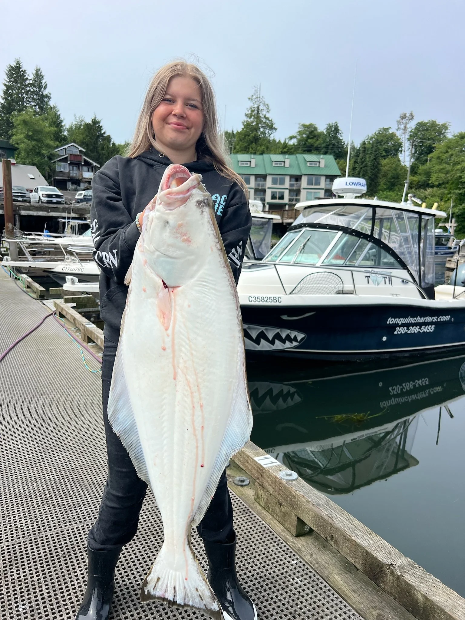 A young woman with long blonde hair in a black hoodie and black boots holding a large fish on a dock at a marina with boats and green trees in the background.