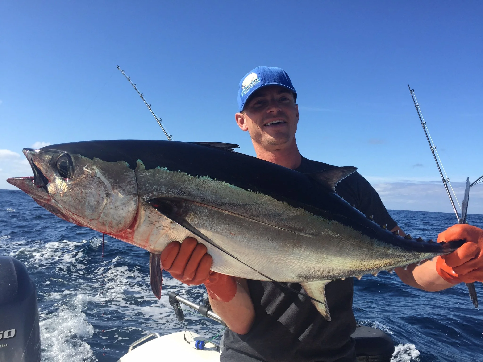 A man wearing a blue cap and black shirt holding a large fish on a boat, with fishing poles and ocean in the background.