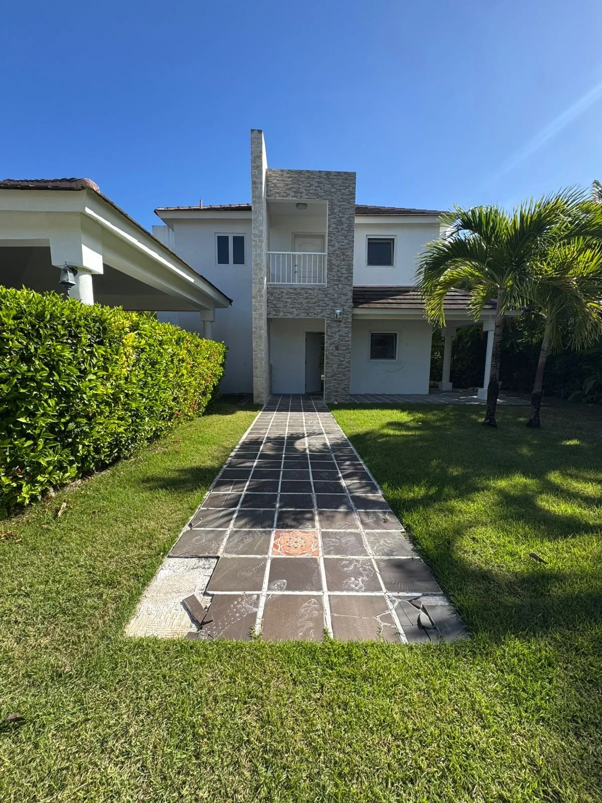 Front view of a modern white house with a tiled pathway leading to the front door, green bushes on the left, palm trees on the right, and a clear blue sky above.