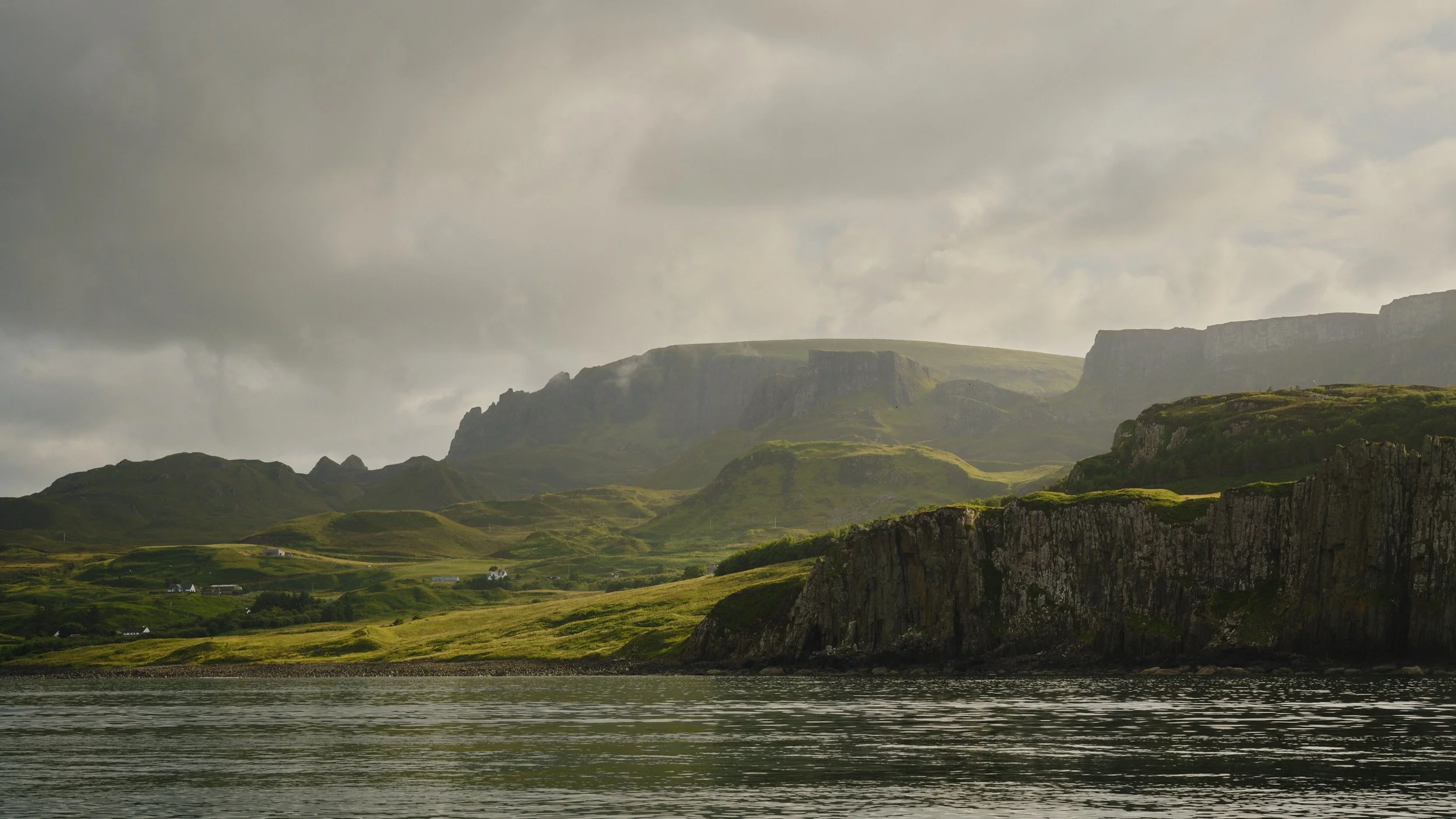 A landscape of green rolling hills with a few scattered houses, tall cliffs in the foreground, and mountains in the background under a cloudy sky.