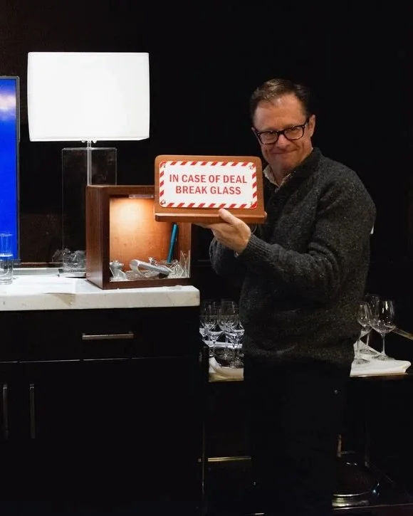 A man holding a box with a sign that reads "In Case of Deal Break Glass" in a kitchen or bar area, with glasses on the counter behind him.