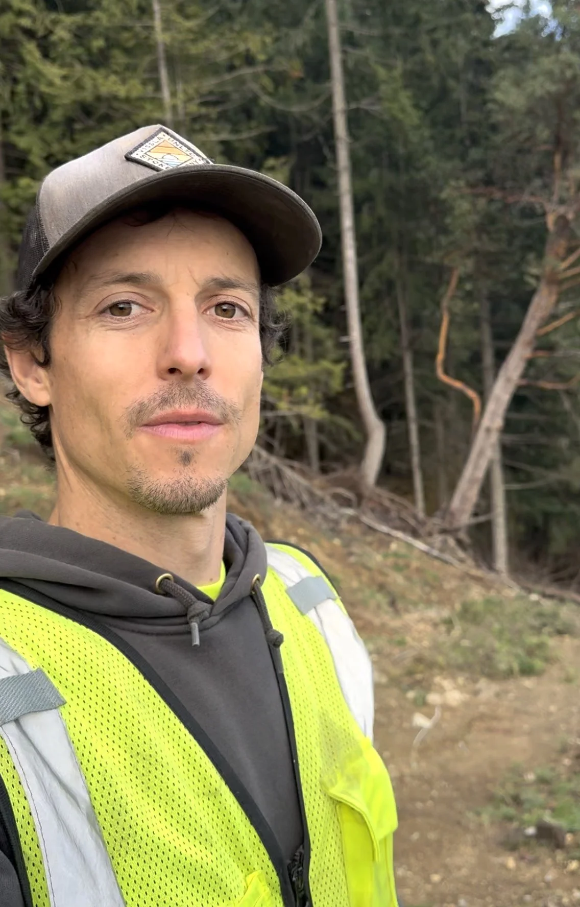A man wearing a gray cap, black hoodie, and yellow safety vest standing outdoors in a wooded area.