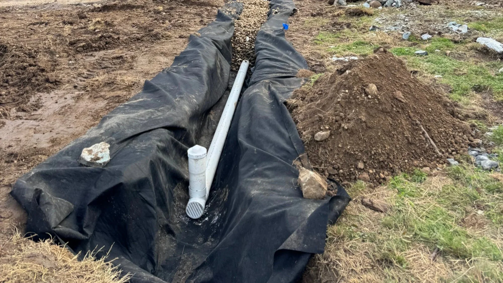 An outdoor construction scene showing a trench covered with black plastic sheeting. A white PVC pipe runs through the trench, with rocks placed along the pipe and on the ground. Soil and grass are visible around the trench.
