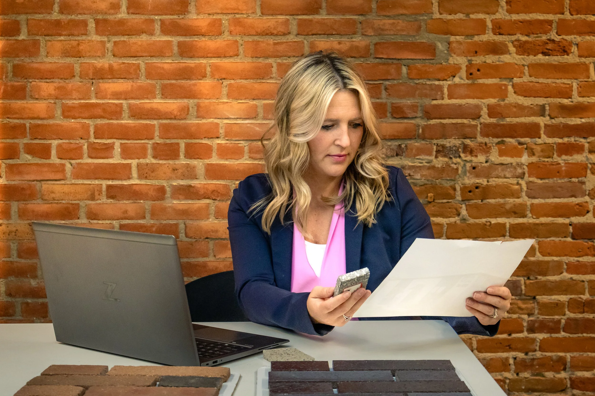 Leah Goodman of E.H. Goodman Architects. A woman with blonde hair sitting at a table in front of a brick wall, examining a piece of paper she is holding. There is a gray laptop and samples of brick colors on the table.