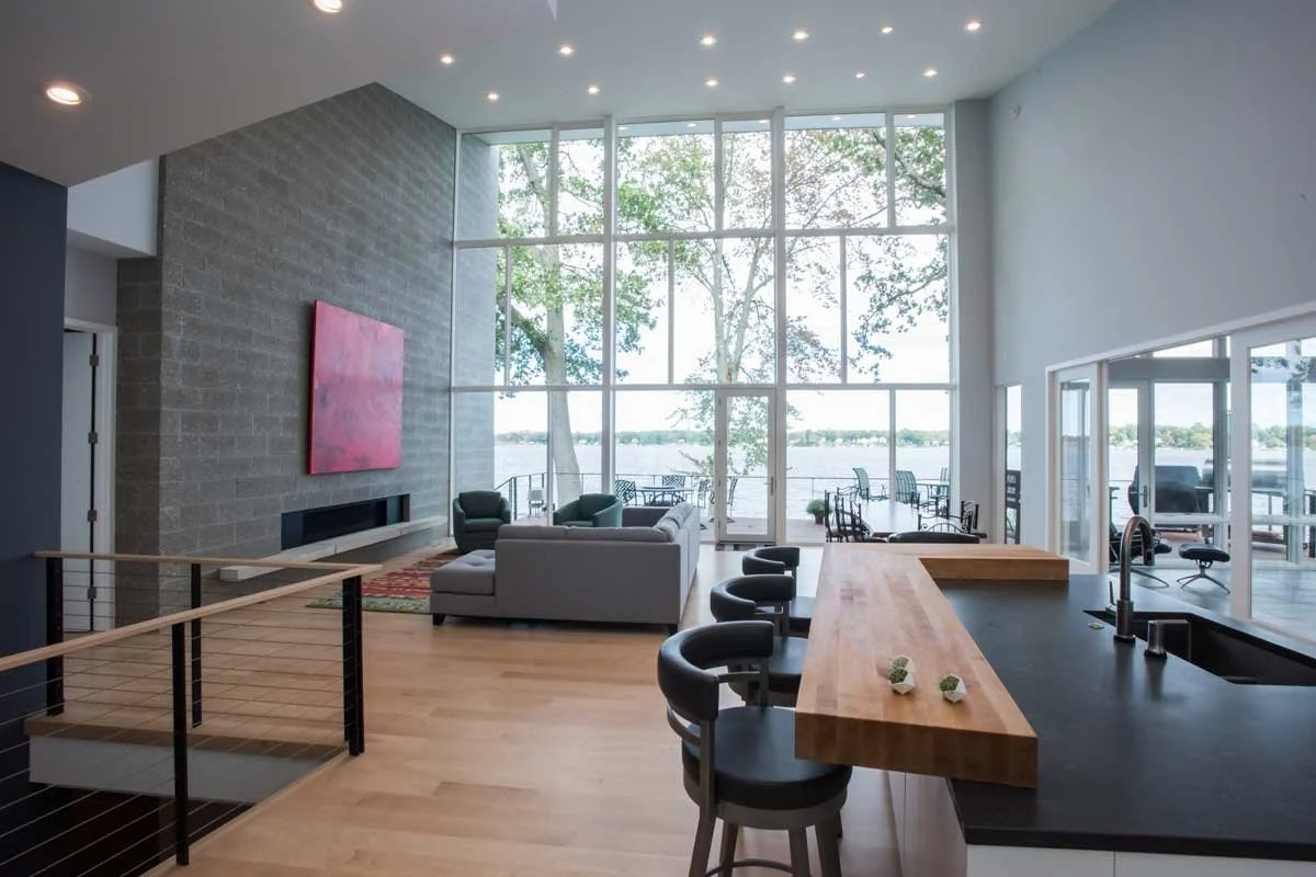 Modern living room design with large floor-to-ceiling windows overlooking a lake, with a seating area, fireplace, and a bar counter with bar stools.