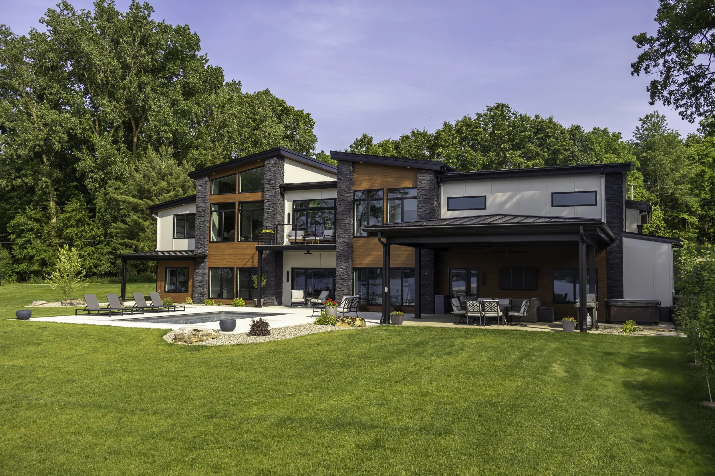 Modern house design with large windows, outdoor patio, pool, lush green lawn, and trees in the background under a clear blue sky.