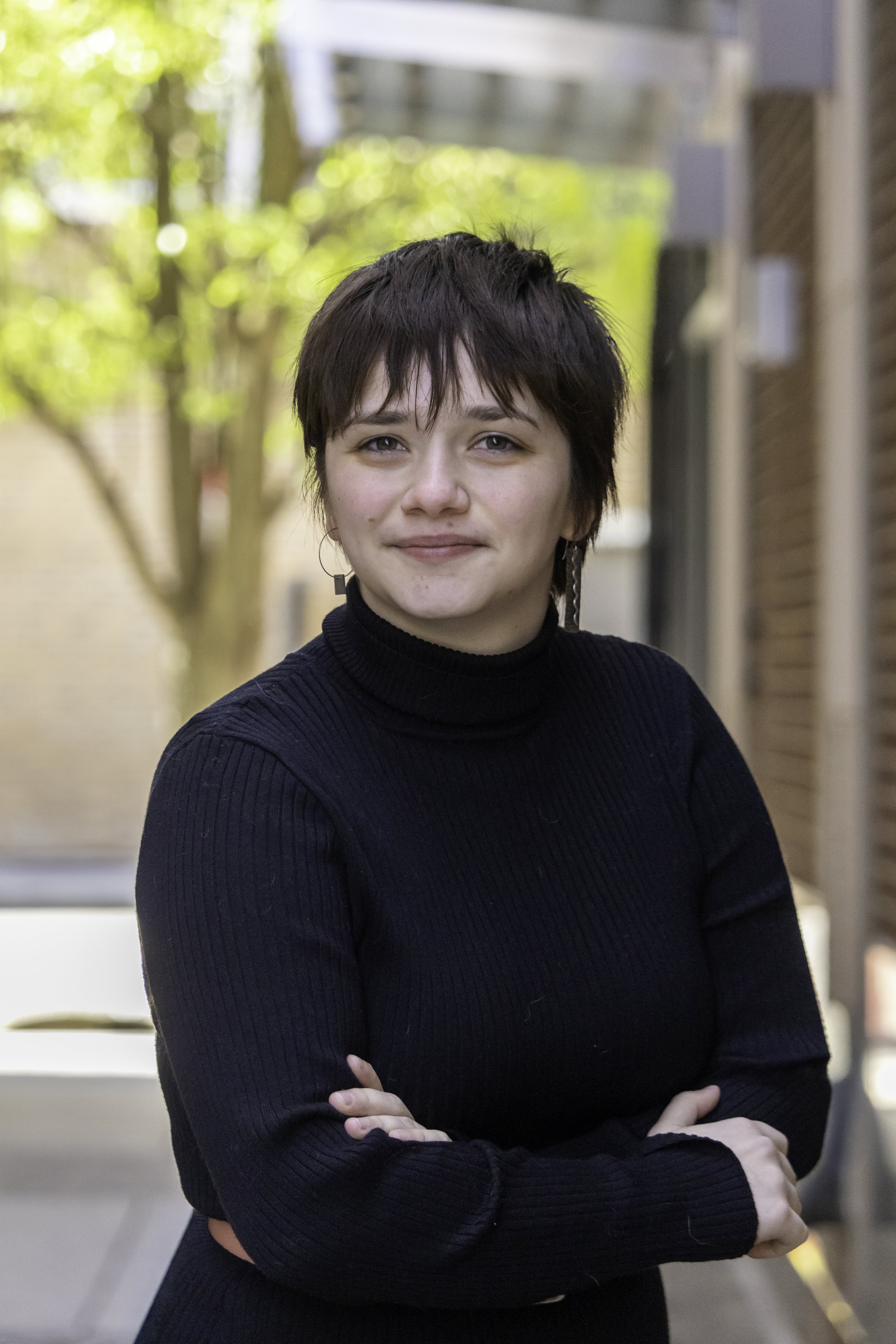 Mali Jean Borisch, Architectural Intern of E.H. Goodman Architects. A young woman with short dark hair, wearing a black ribbed turtleneck sweater, standing outdoors with trees and building in the background, smiling slightly with her arms crossed.