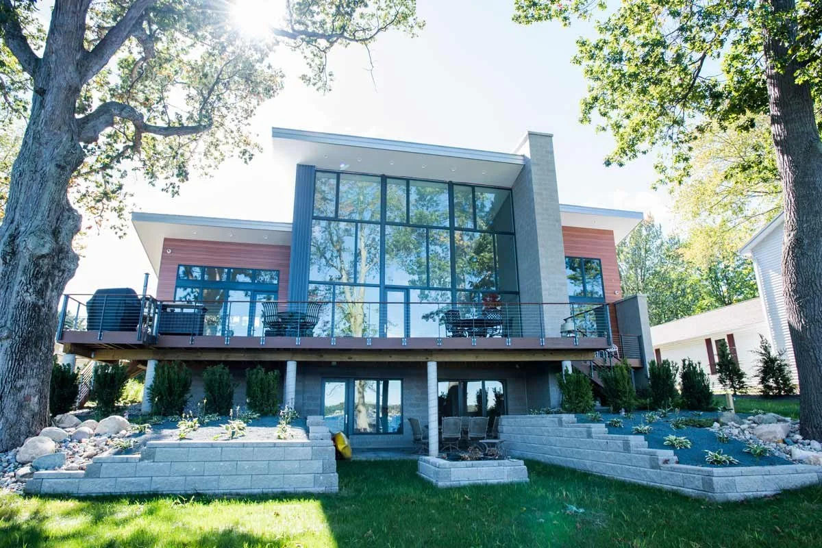 Modern house with large glass windows, a balcony with outdoor furniture, and landscaped yard with trees and garden beds