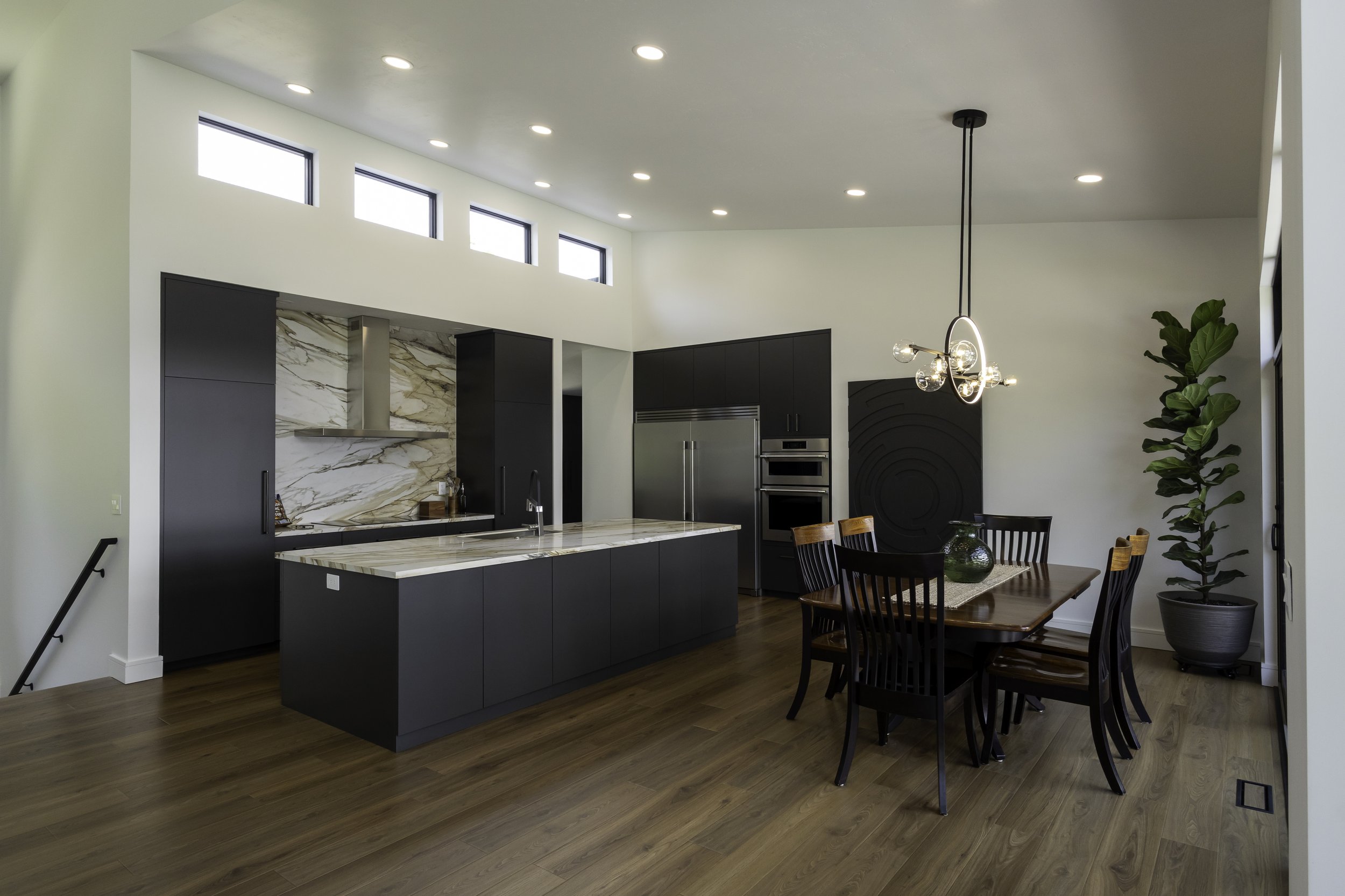 Modern kitchen and dining area with black cabinetry, marble backsplash, pendant lighting, large windows, and a wooden dining table with chairs.
