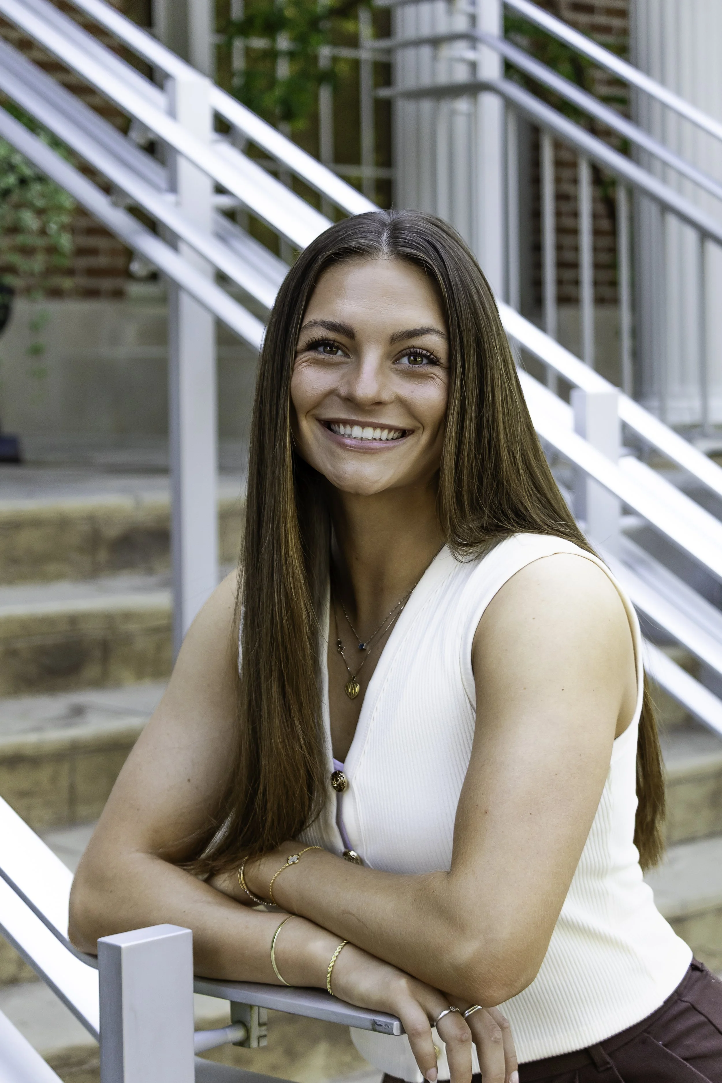 Ashley Bates Architectural Intern of E.H. Goodman Architects. A smiling woman with long brown hair wearing a white sleeveless, standing outdoors on a staircase with metal railings and greenery in the background.