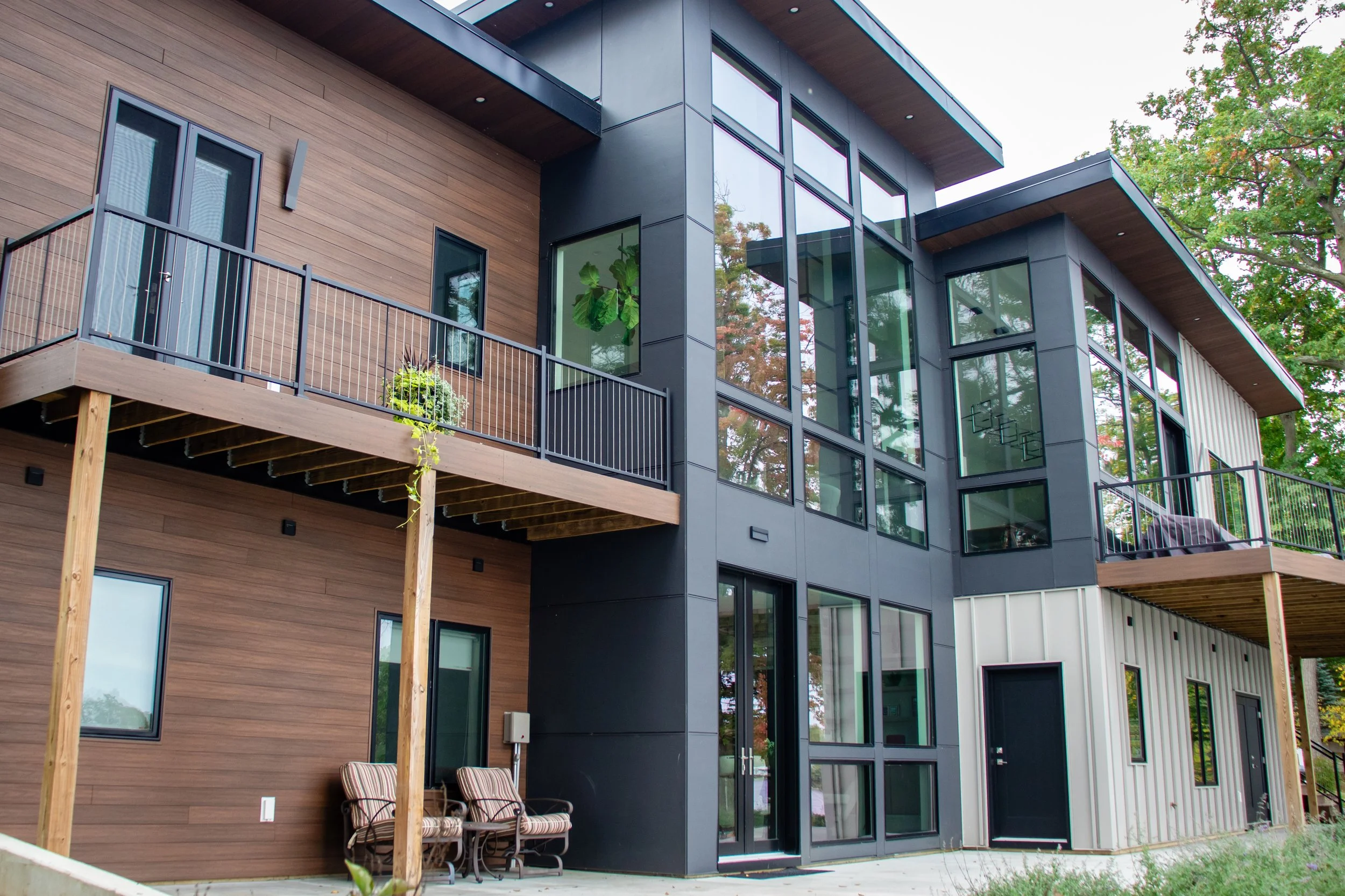 Modern multi-story modern house design with large glass windows, wooden and dark gray exterior, balcony with metal railing, and outdoor seating area with two chairs.
