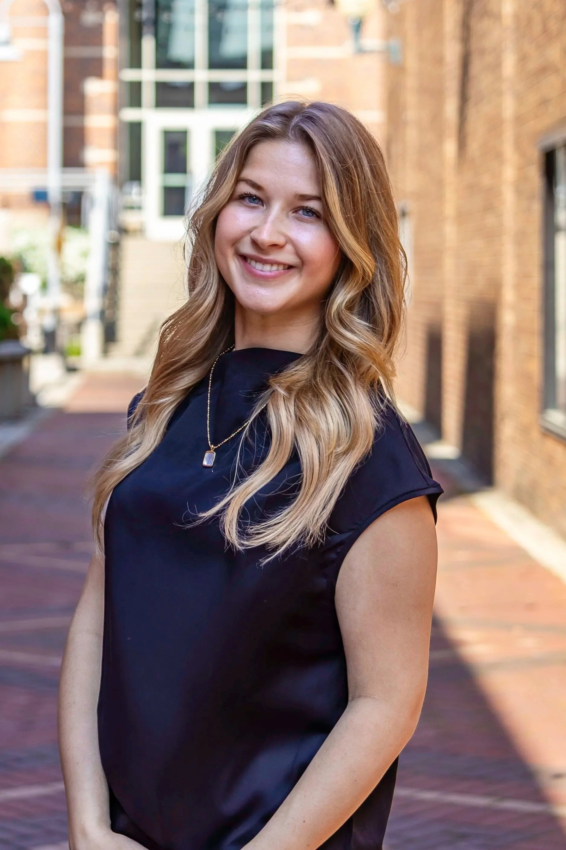 Sarah Fuchsgruber Interior Design Intern of E.H. Goodman Architects. A young woman with long, wavy blonde hair and blue eyes smiling outdoors in front of a brick building.