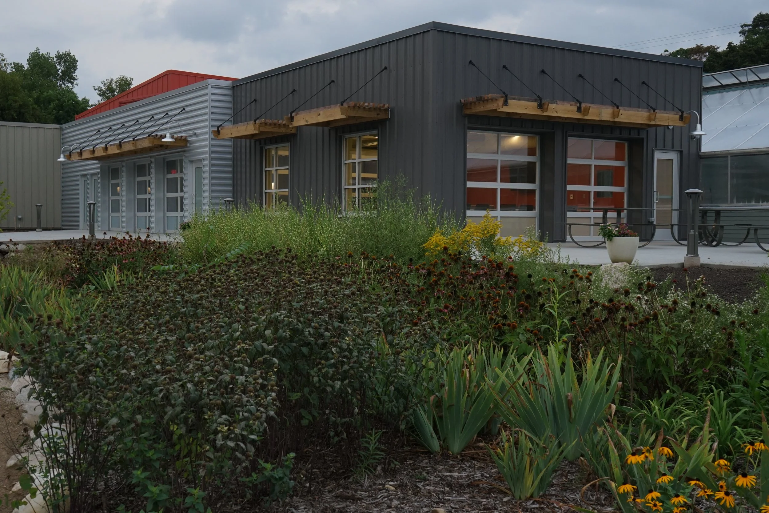 Exterior view of a modern building with a gray and silver metal façade, featuring large windows, outdoor lighting fixtures, and wooden canopy structures. The building is surrounded by lush green plants and flowers in a landscaped garden.