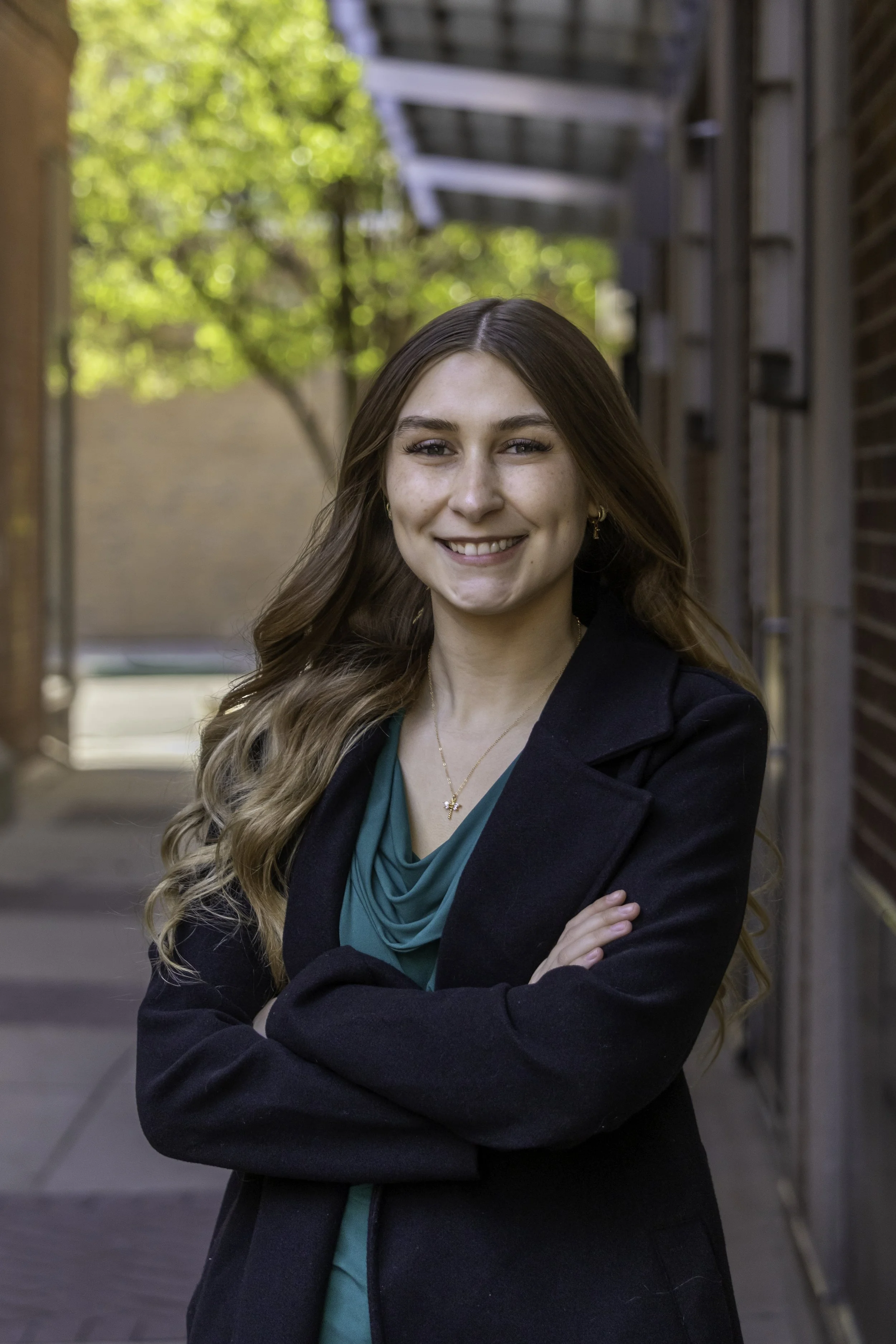 Lillyan Gurd Interior Design Intern of E.H. Goodman Architects. young woman with long wavy brown hair and light skin, smiling and arms crossed, standing outdoors near a brick building with a wooden fence, wearing a black blazer and teal blouse.