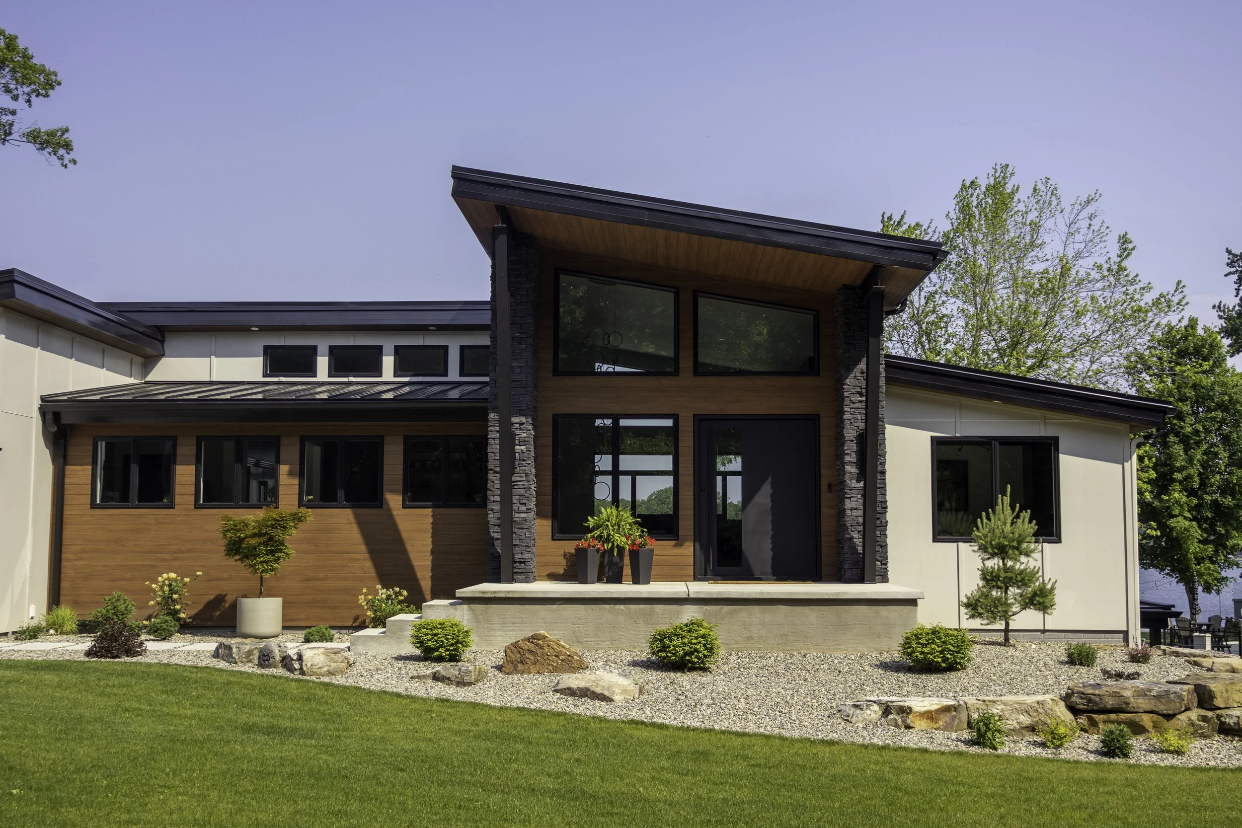 Modern house with large windows, wooden and stone exterior, front porch with potted plants, surrounded by green lawn and landscaping, under a clear blue sky.
Modern House Design in Southwest Michigan.