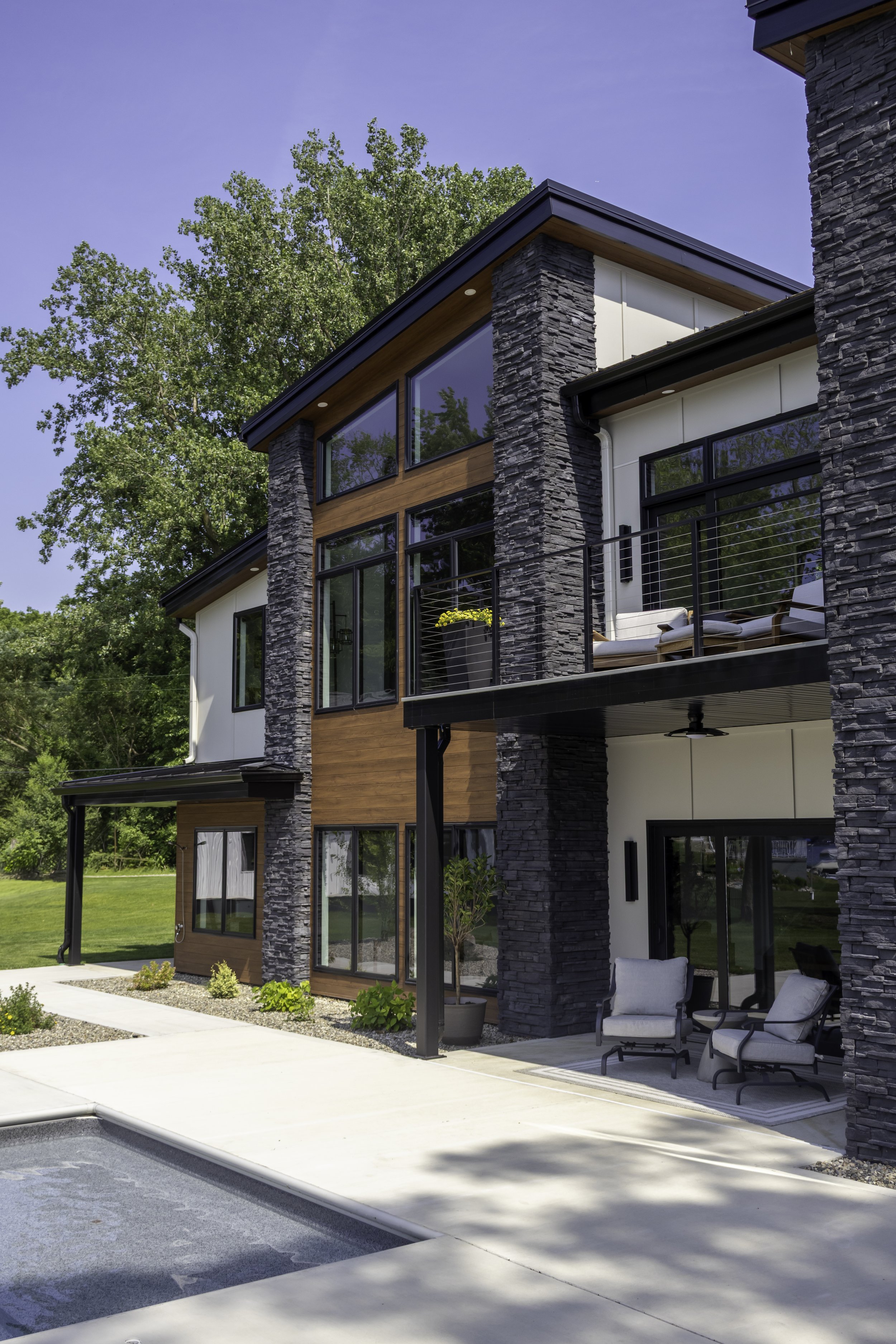Modern house with stone and wood exterior, glass windows, balcony, and outdoor seating area, surrounded by greenery under a clear blue sky.
Luxury Modern house design.