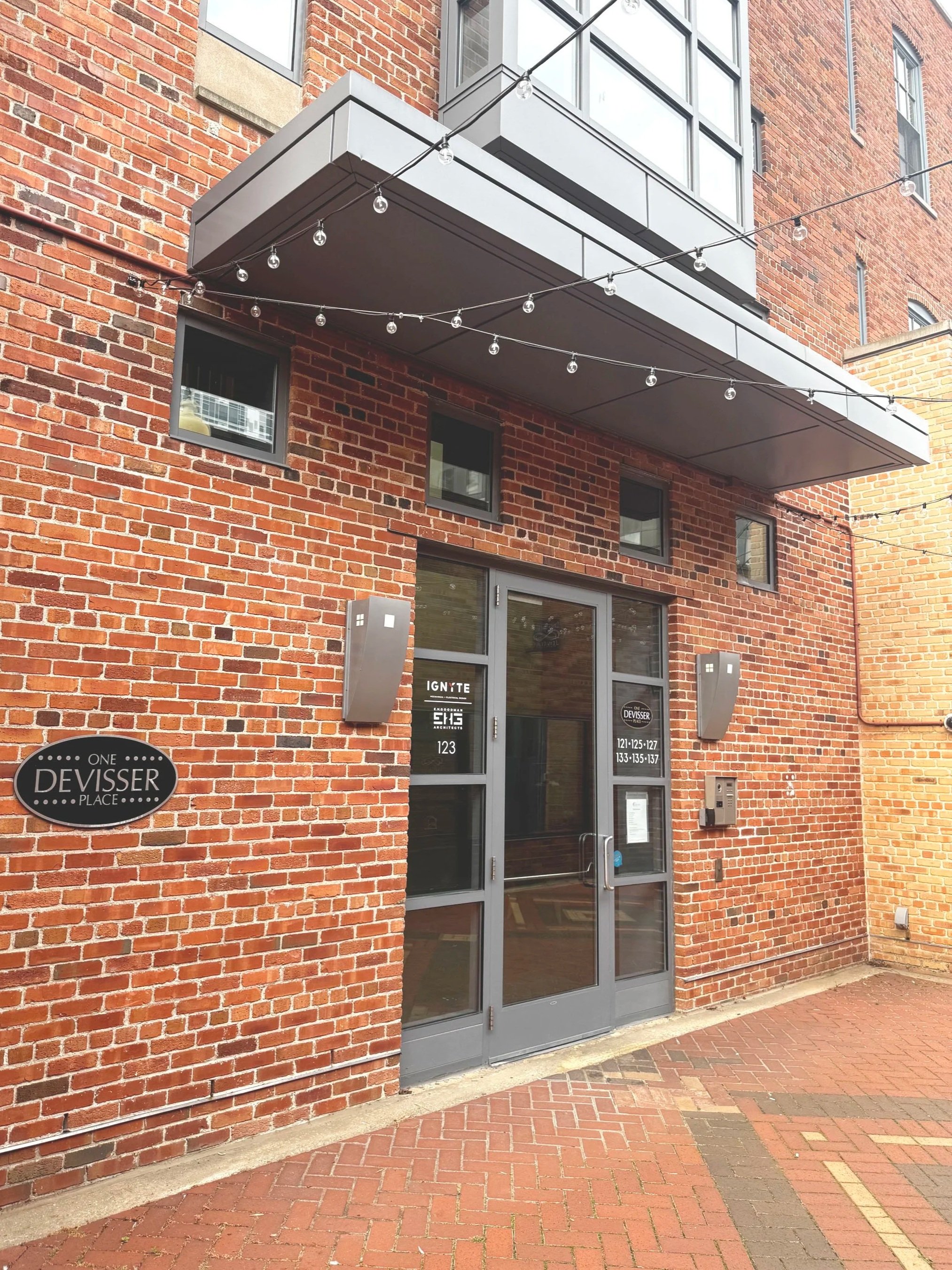 The entrance of a brick building with a glass door, two wall-mounted mailboxes with the sign 'ONE DEVISSER PLACE,' and a street address number '123.' String lights are hanging above the entrance.