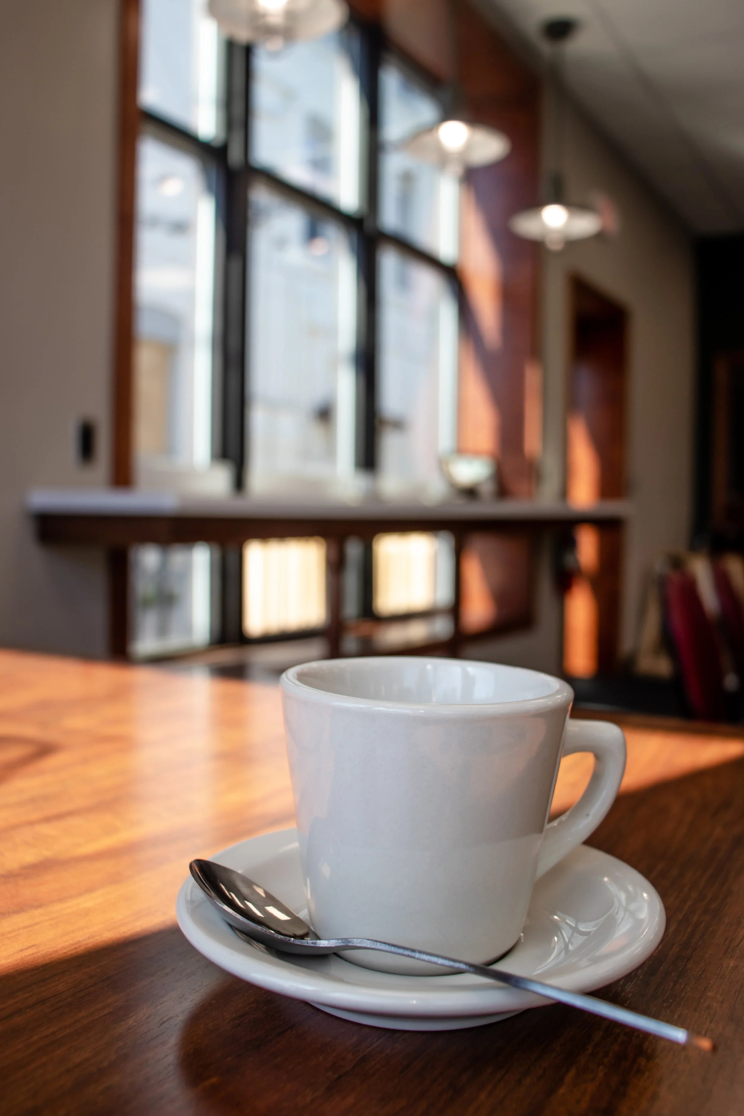 White coffee cup with a spoon on a saucer on a wooden table in a coffee shop, with sunlight streaming through large windows in the background.
KPep Coffee Shop Renovation.