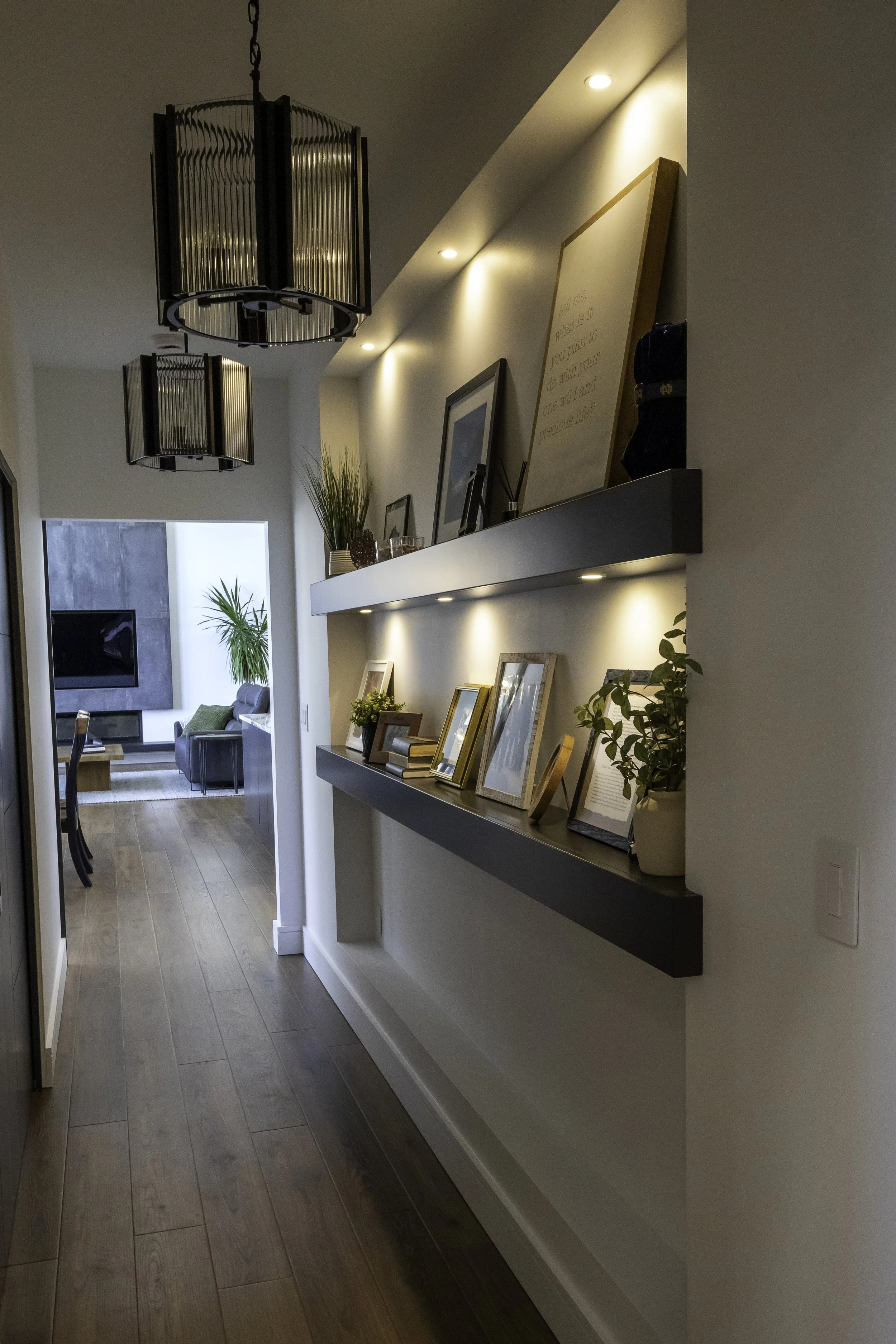 Interior of a modern home with a hallway leading to a living room, features include a floating shelf with framed photos, plants, and artwork, ceiling lights, and wooden flooring.