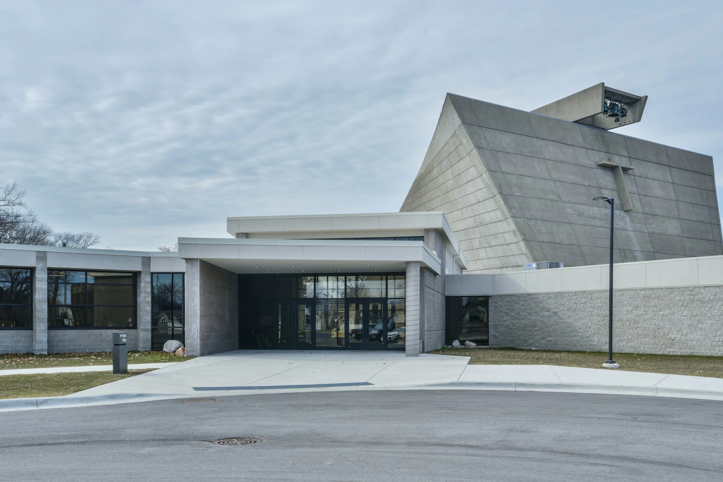 A modern church building design with stone and glass exterior, featuring angular concrete architecture and an overhang with lights, against an overcast sky.
