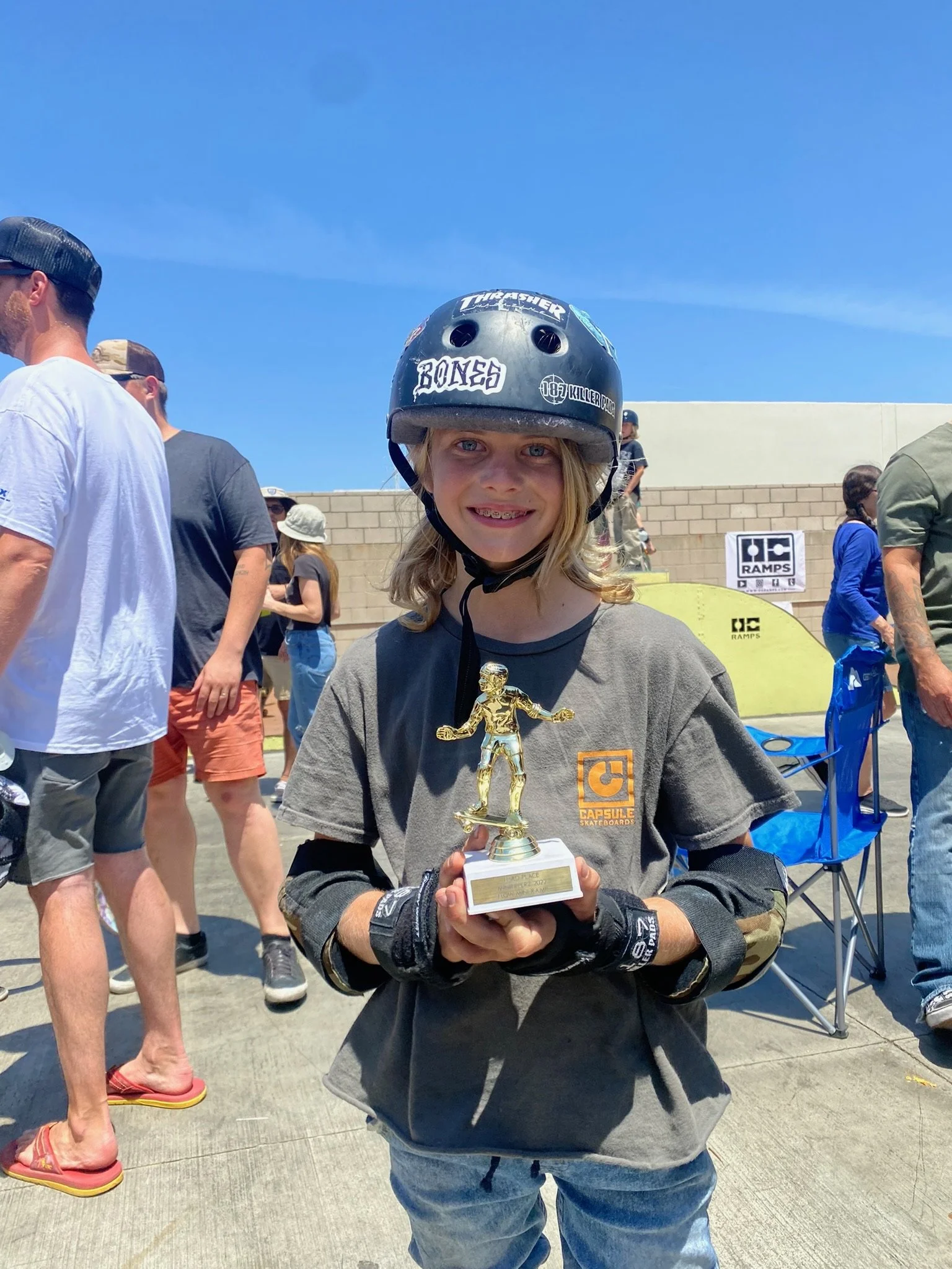 A young girl with a helmet and protective gear smiling and holding a trophy, surrounded by people outdoors on a sunny day.