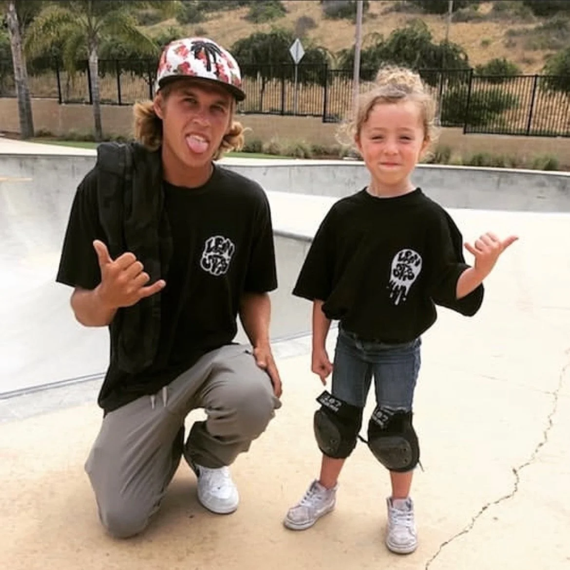 A teenage boy and a young girl at a skatepark, both wearing black T-shirts with a logo, making hand gestures, and smiling or smirking. The boy is kneeling with one knee on the ground, and the girl is standing with knee pads on.