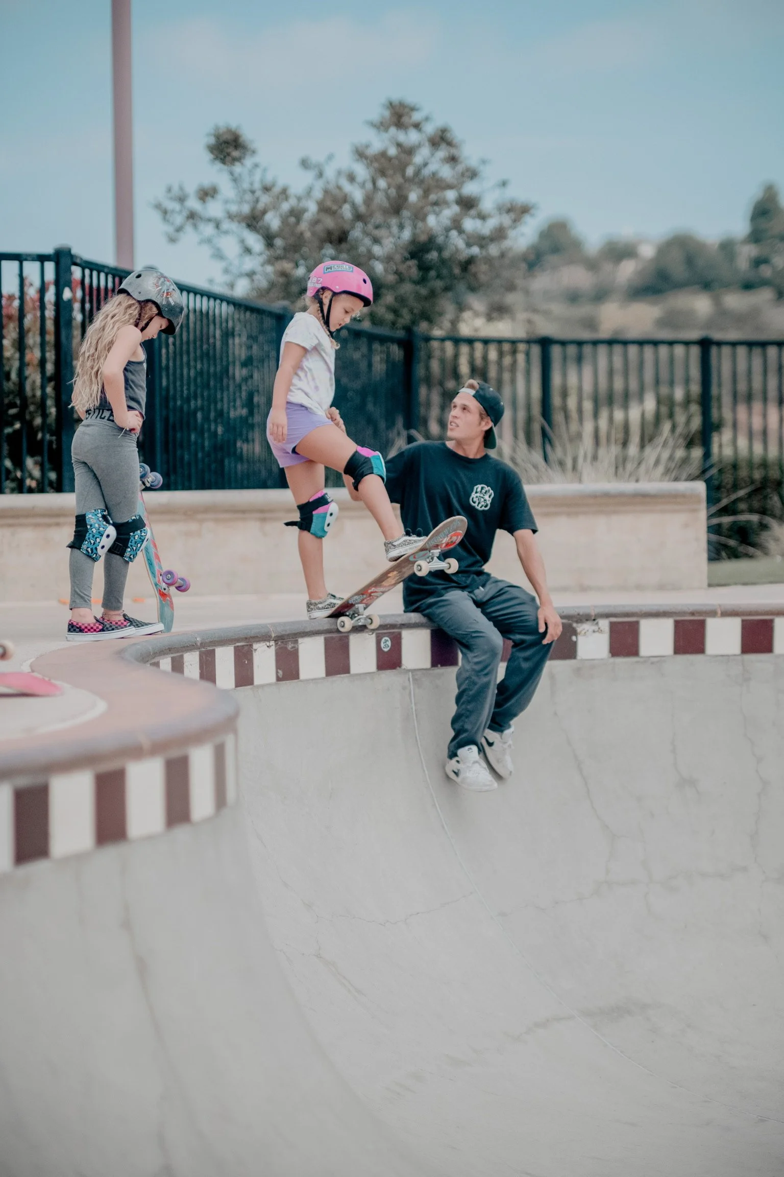 Three children at a skate park, with one child riding a skateboard and two children watching, all wearing helmets.