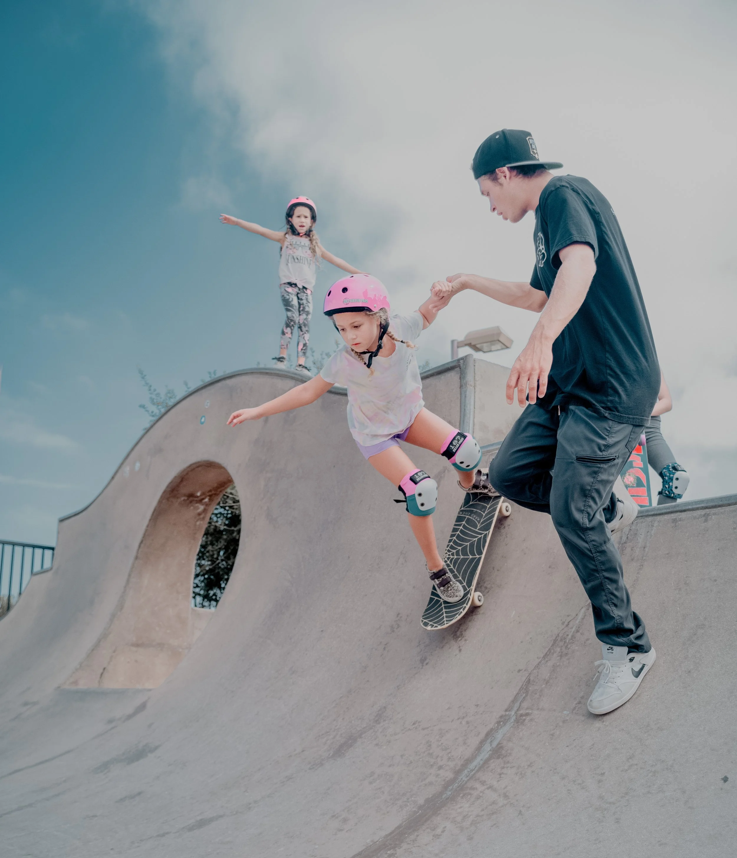 Two young girls skateboarding at a skate park with a young man assisting, under a cloudy sky.