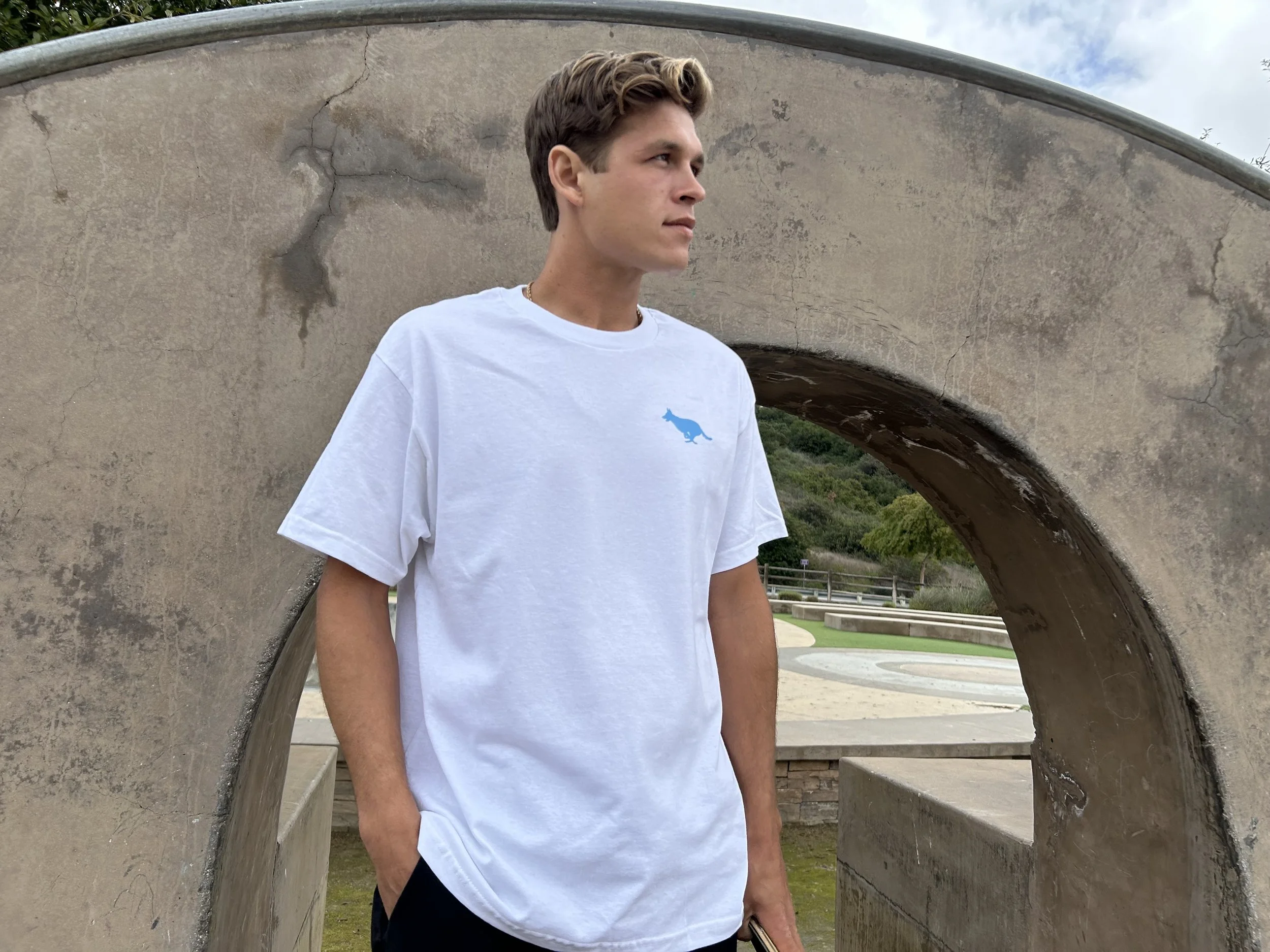 A young man with short, curly brown hair gazes to the right, standing near a large, weathered concrete arch with visible cracks. He is wearing a white T-shirt with a small blue dinosaur logo on the chest and a gold chain necklace. The background features greenery and a partly cloudy sky.