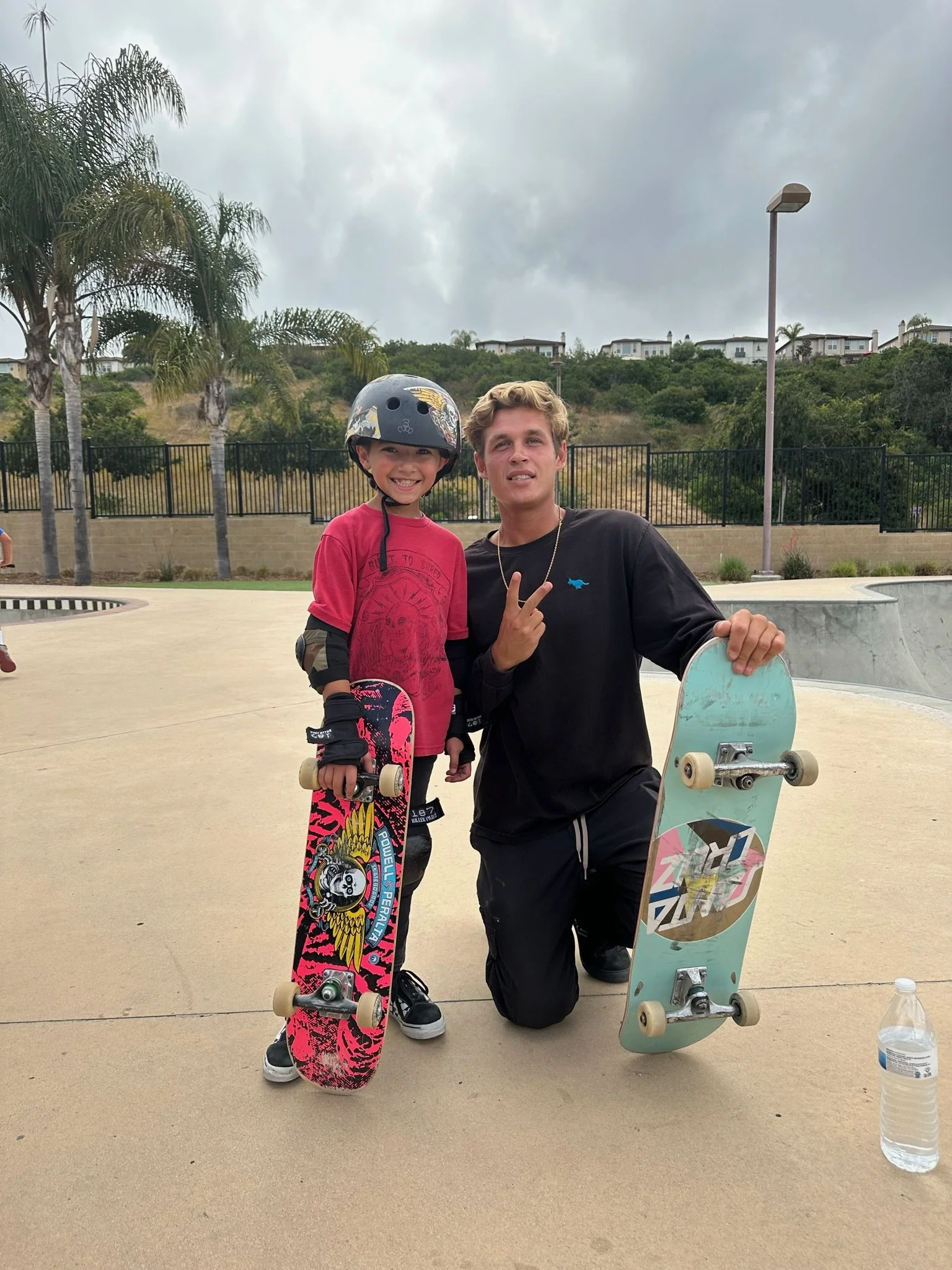 A smiling young boy wearing a helmet and protective gear holding a colorful skateboard, posing beside a man kneeling with a skateboard at a skate park on a cloudy day, with palm trees, a fence, and residential houses in the background.