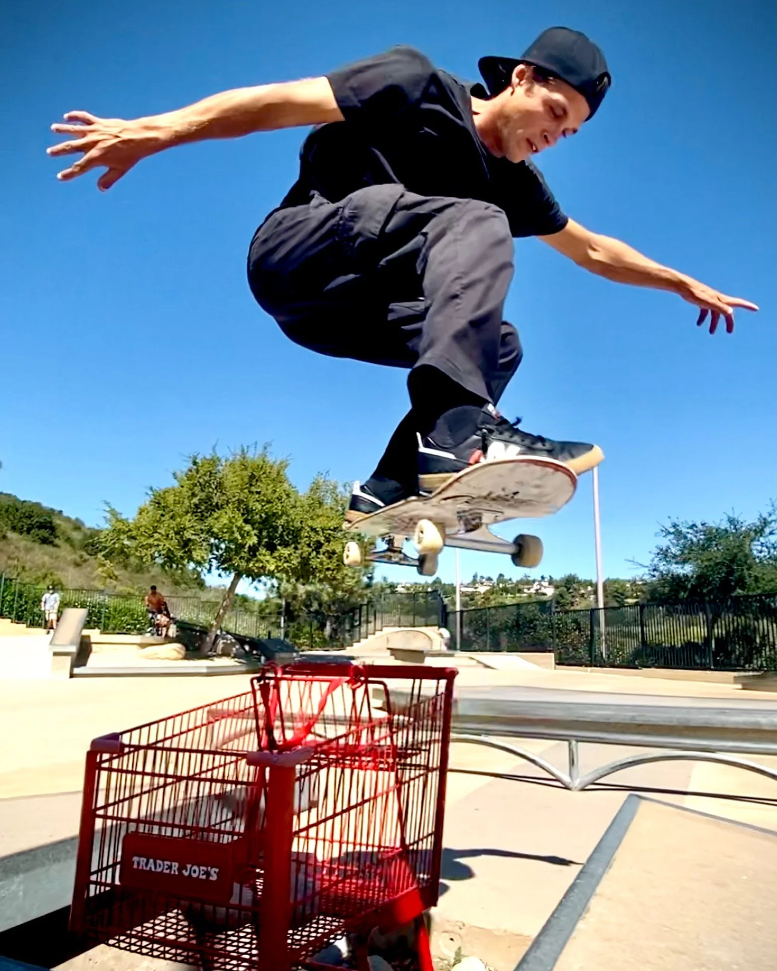 A person skateboarding outdoors on a sunny day with trees and a clear blue sky in the background, viewed from ground level with a red shopping cart in the foreground.