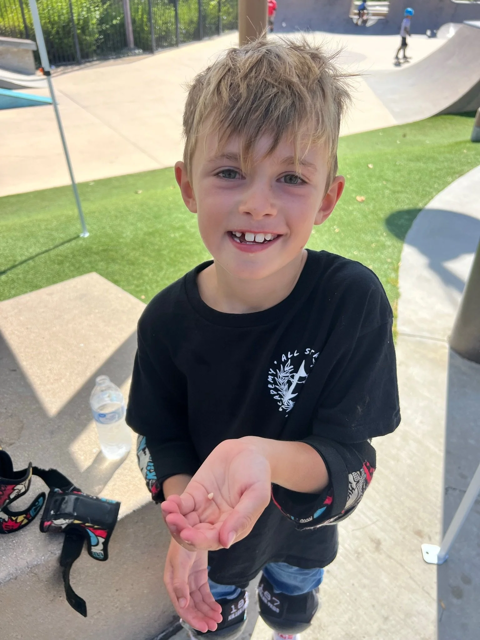 A young boy smiling and extending his hand toward the camera at an outdoor skate park, wearing a black T-shirt and rollerblades.