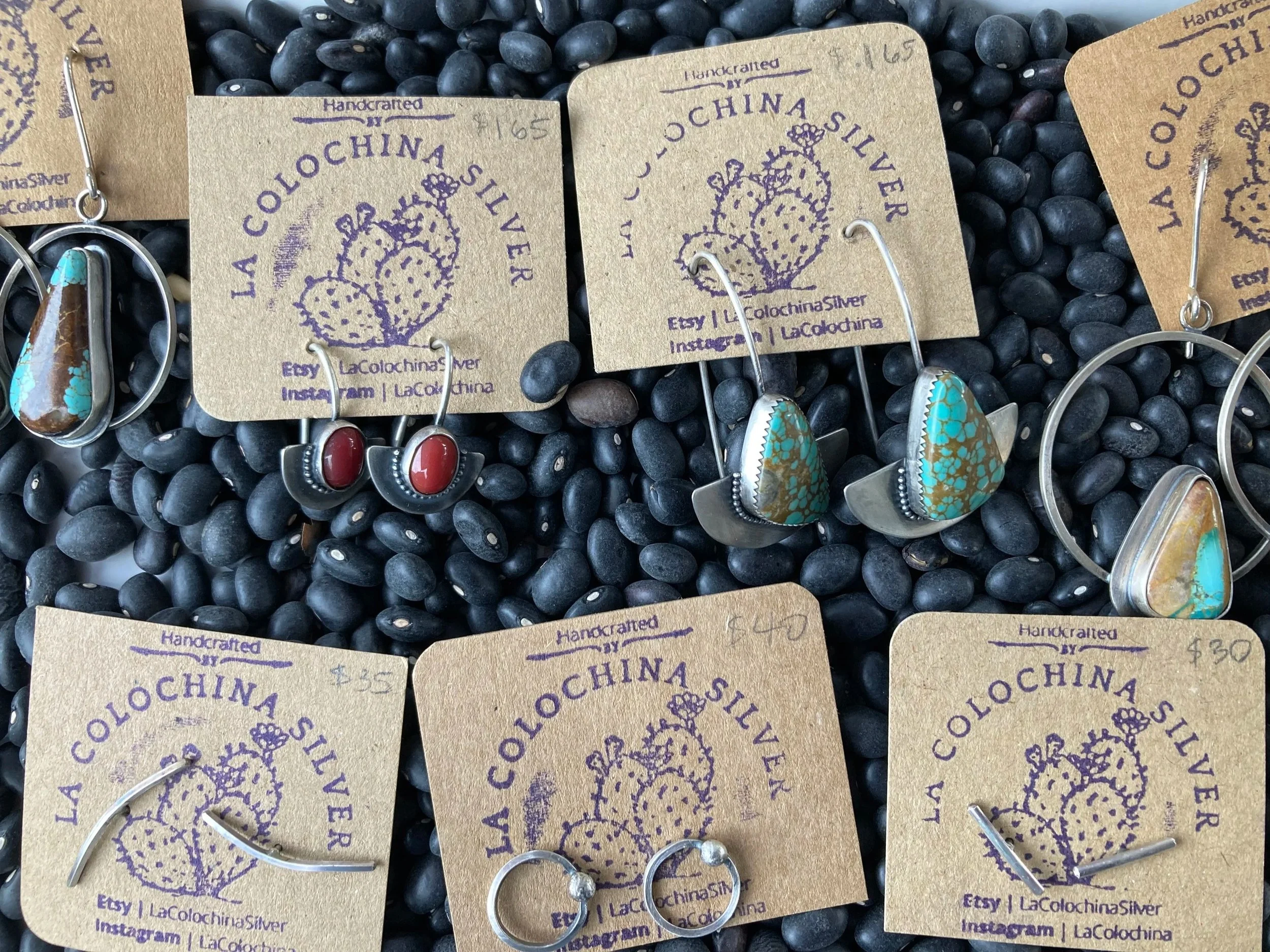 Close-up photo of handcrafted earrings on brown cards with purple text and logo, displayed on black stones. The cards read "Handcrafted by LaColochina Silver," with the website and Instagram handle. The earrings feature turquoise and red stones with silver settings.