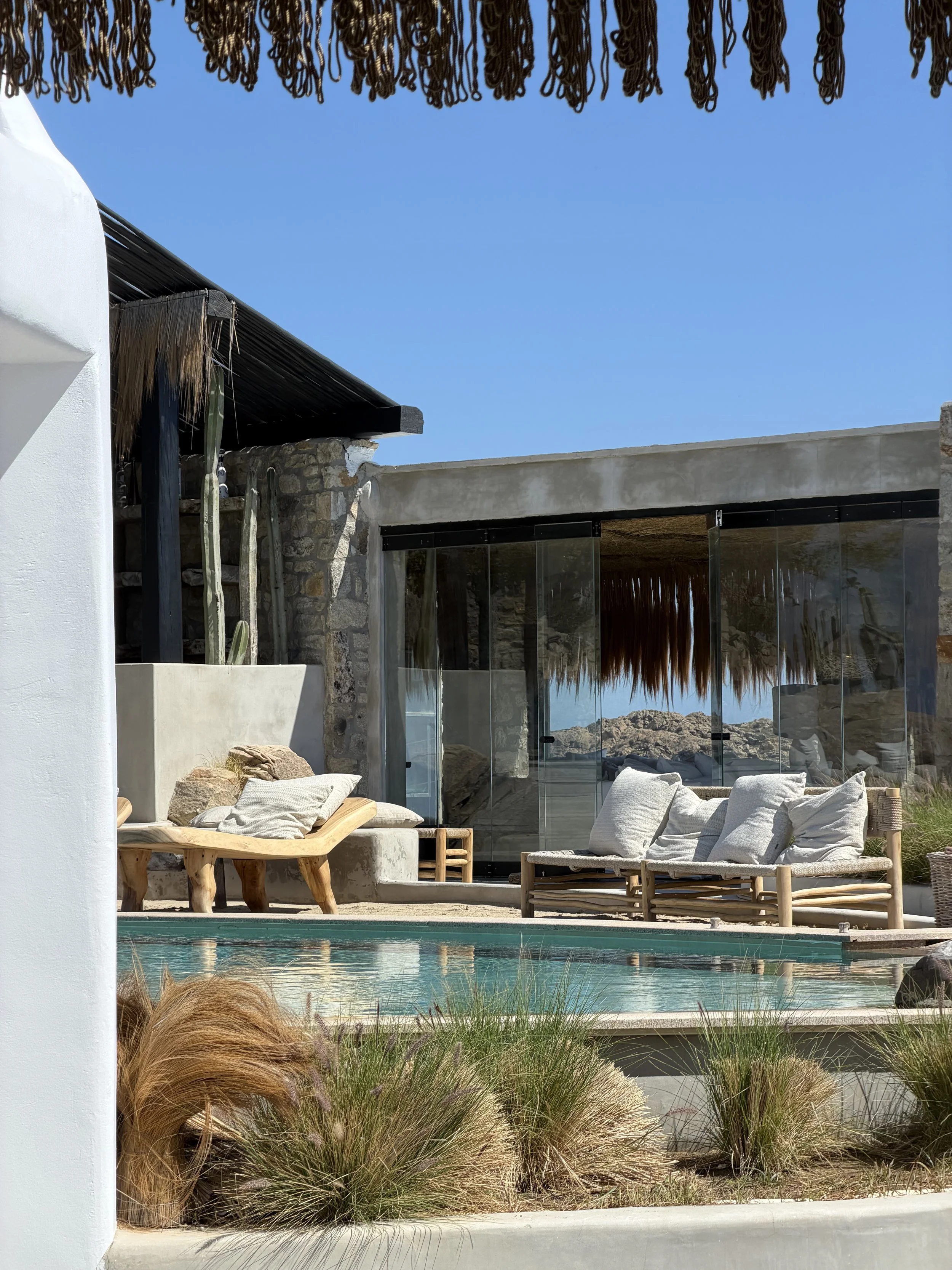 Ein moderner Außenpoolbereich mit Liegestühlen, einem Gartengrundstück mit Gräsern und Pflanzen, einer Villa mit Glaswänden und einer Dachterrasse, im Hintergrund Felsen und klare blauer Himmel.