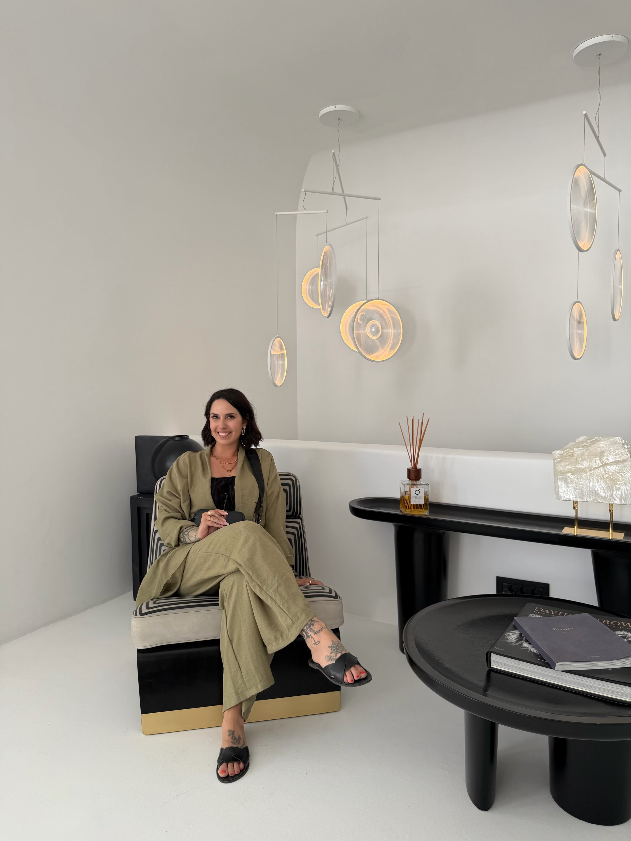Eine lächelnde Frau sitzt entspannt auf einem bequemen Stuhl in einem modernen, minimalistischen Raum mit weißen Wänden und eleganter Beleuchtung. Sie trägt einen grünen Anzug und schwarze Sandalen. Auf einem schwarzen Beistelltisch liegt ein Buch, daneben befindet sich eine Dekoration und eine Rakete.