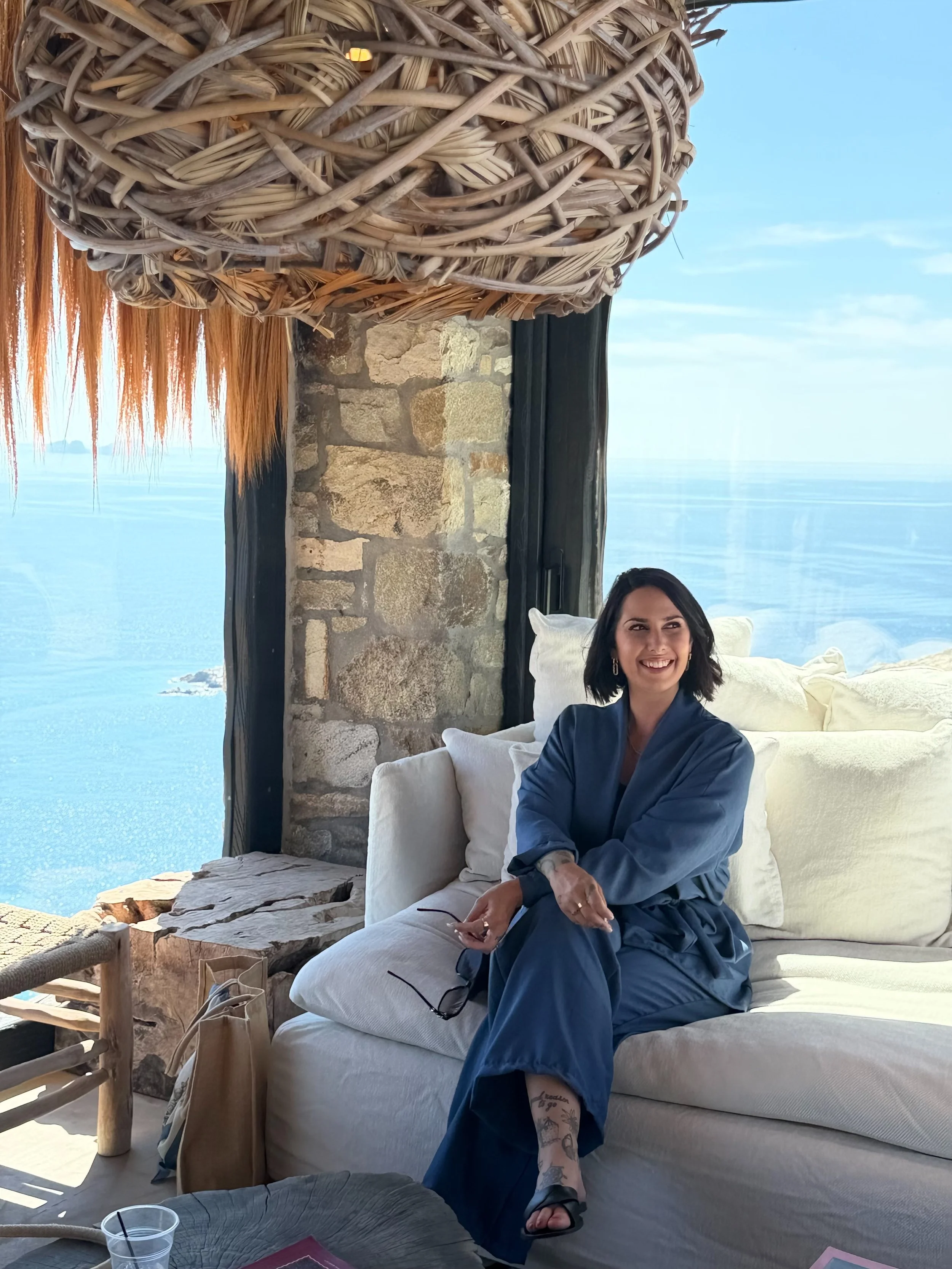 Eine lachende Frau mit dunklem Haar sitzt lächelnd in einem Strandrestaurant mit Blick auf das Meer und den Himmel. Sie trägt eine dunkle Kleidungsstück und zeigt tattoos auf ihrem Bein. Im Vordergrund sind ein Stein-Hocker und ein Glas Wasser sichtbar, im Hintergrund eine stone Mauer und eine Muschel-Dekoration.