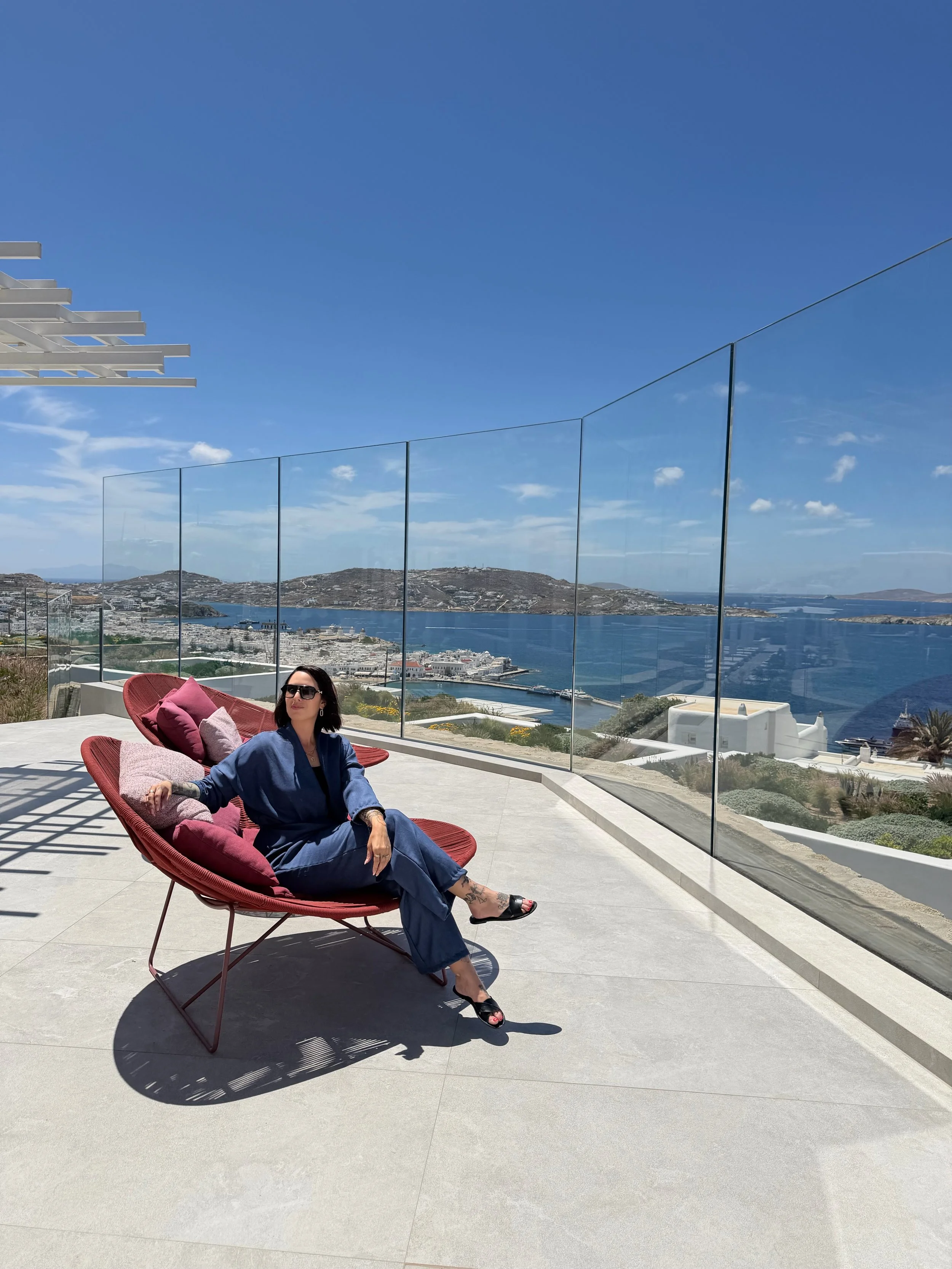 Frau sitzt entspannt in rotem Lounge-Sessel auf einer Terrasse mit Blick auf das Meer und die Hügel im Hintergrund, bei sonnigem Himmel.
