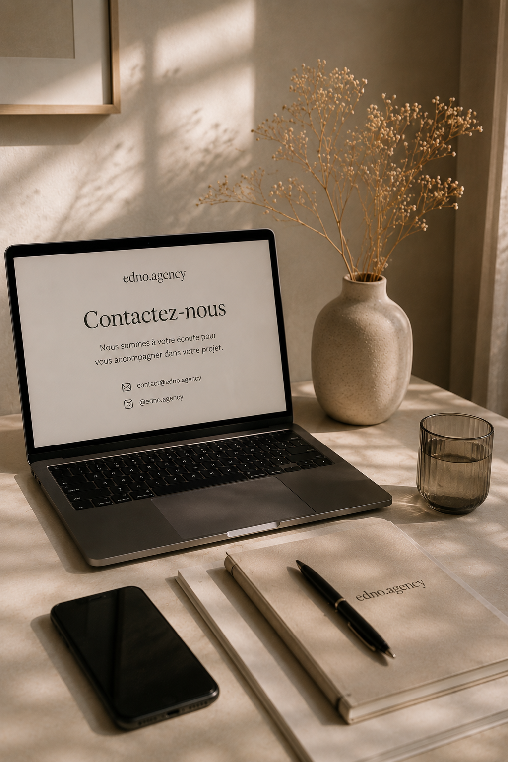 Un bureau avec un ordinateur portable affichant une page de contact, une vase avec des branches séchées, un verre, un téléphone portable noir, un carnet et un stylo.