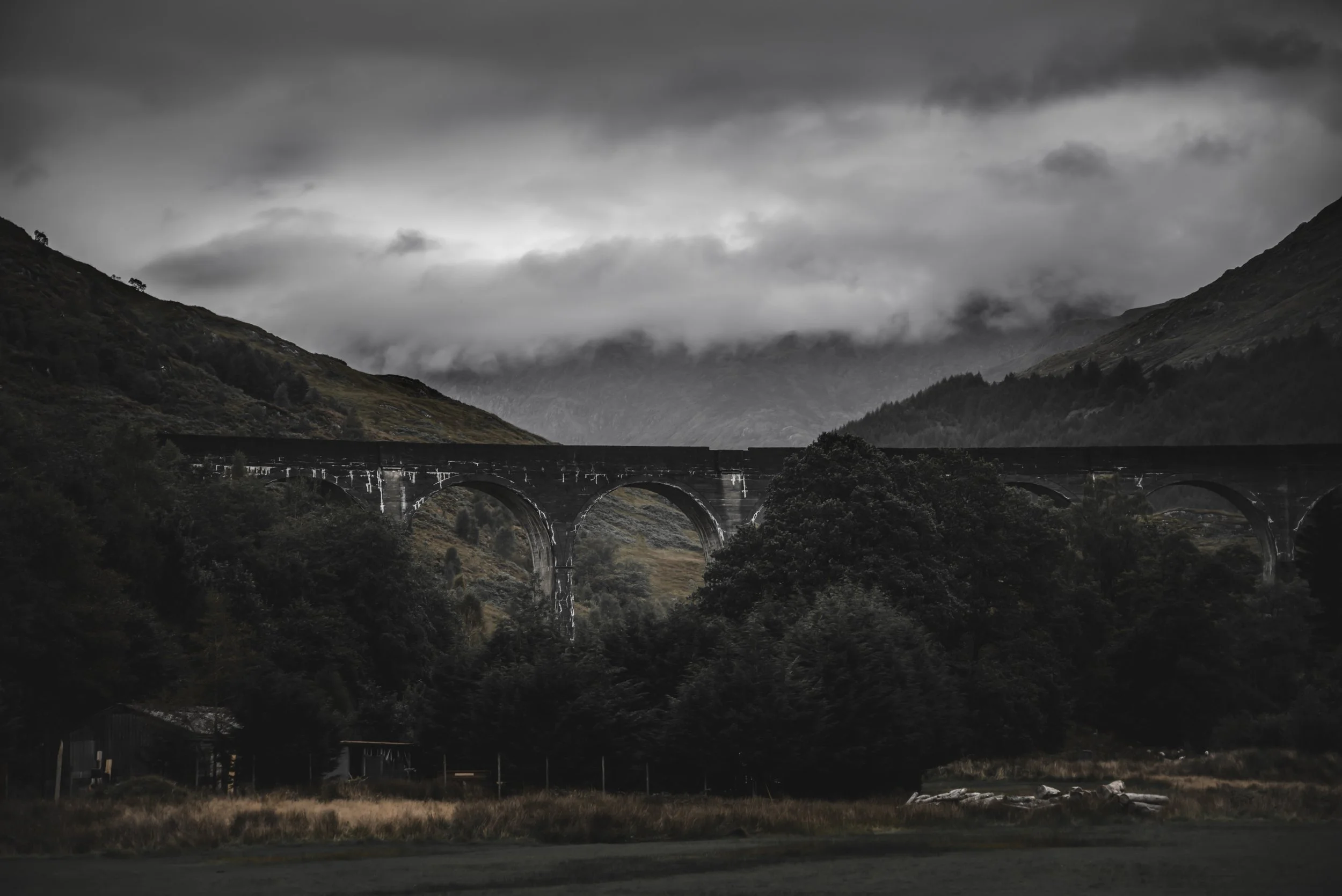 A black-and-white landscape featuring mountains, dark clouds, a stone arch bridge, and trees in the foreground.